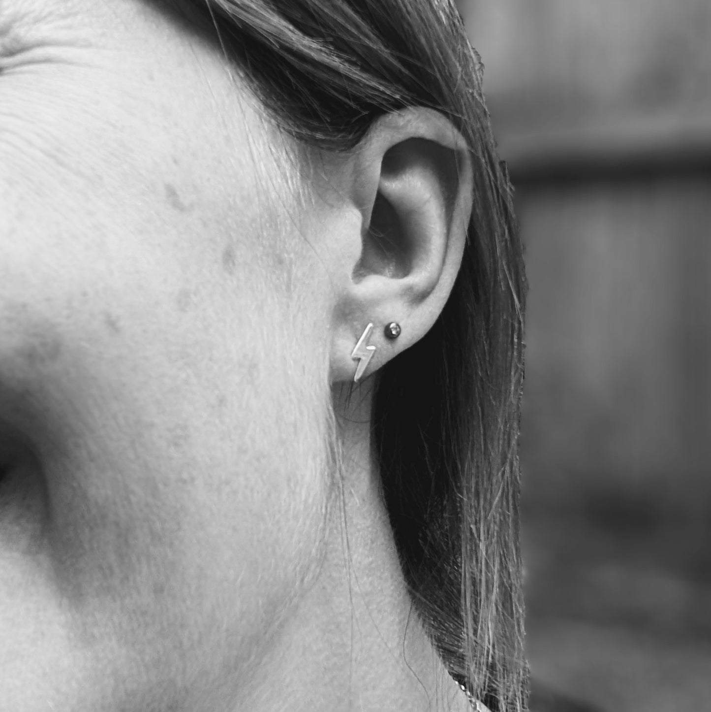 Black and white image of a person wearing a small silver lightning bolt stud earring, shown in the earlobe for size reference.