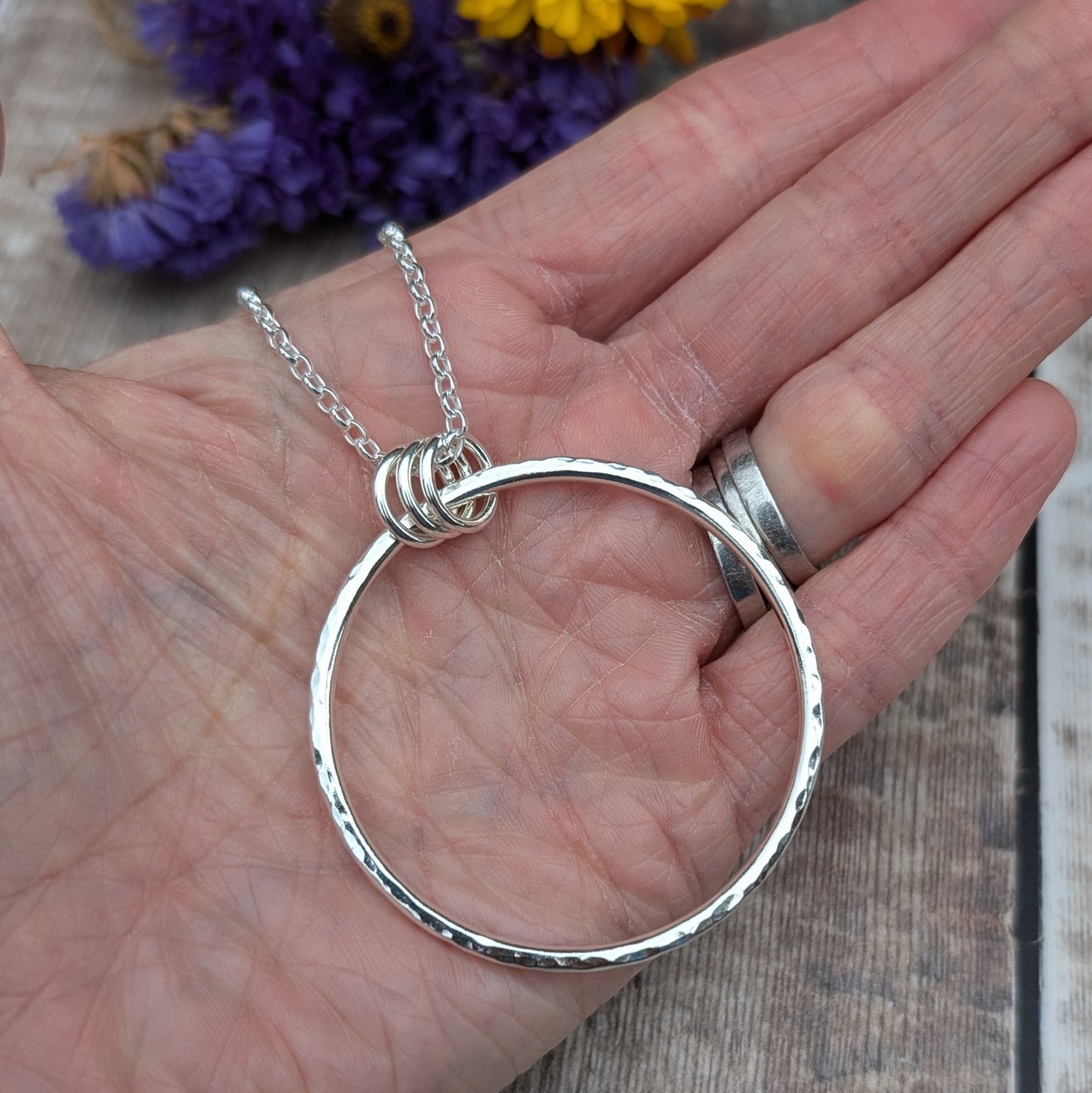 A close-up of a hand holding a large silver hammered circle pendant with three small silver loops at the top, hanging on a silver chain. The background features purple and yellow dried flowers.