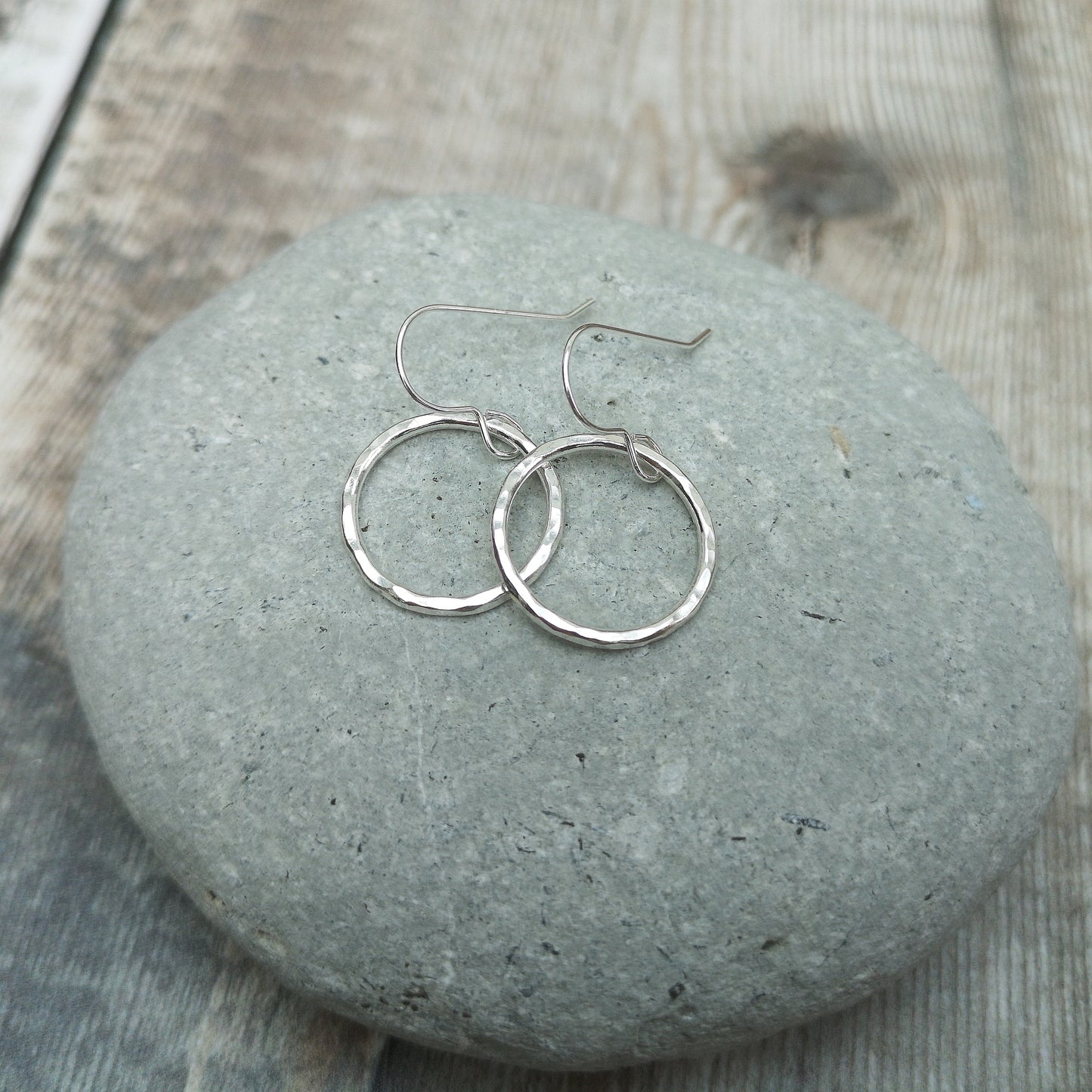 A close-up of medium-sized sterling silver hammered circle earrings resting on a smooth grey stone. The subtle texture of the circles reflects light, enhancing their minimalist and handcrafted design.
