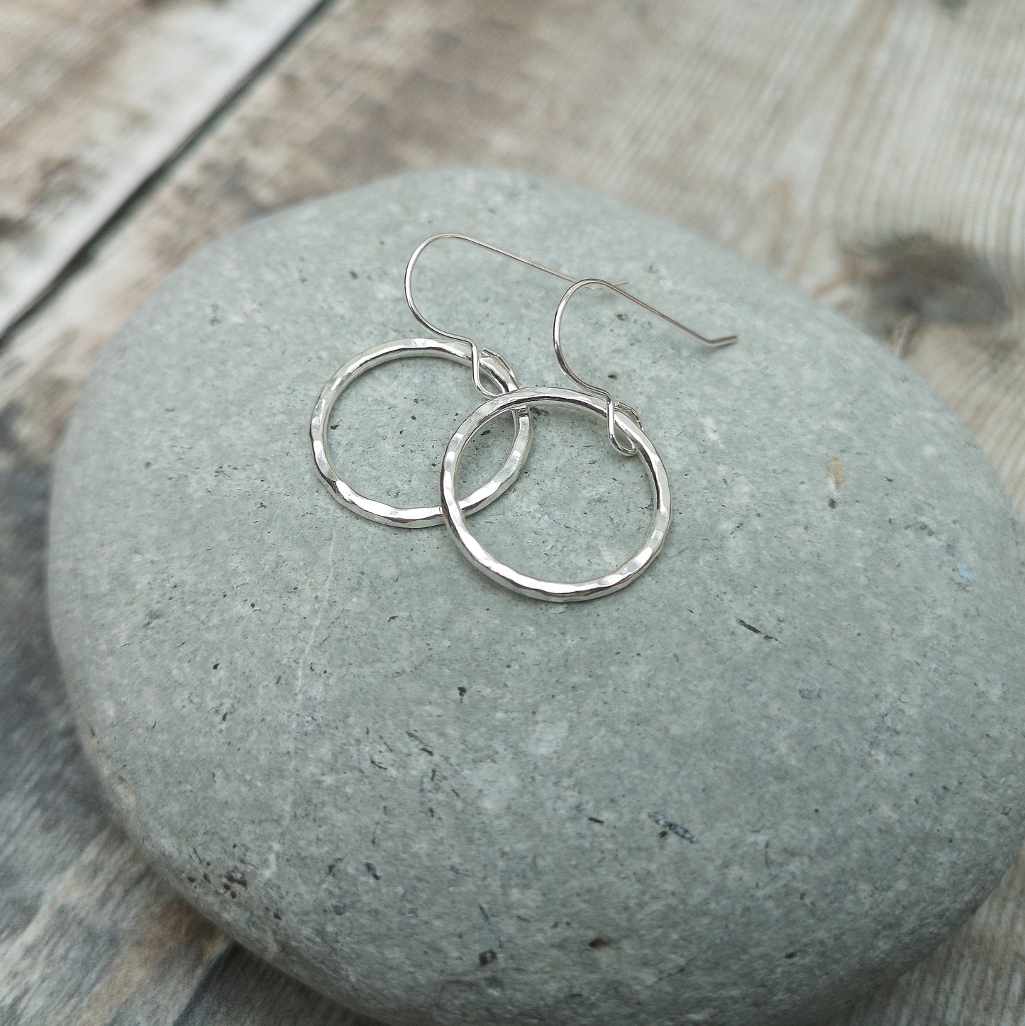 A close-up of medium-sized sterling silver hammered circle earrings resting on a smooth grey stone. The subtle texture of the circles reflects light, enhancing their minimalist and handcrafted design.