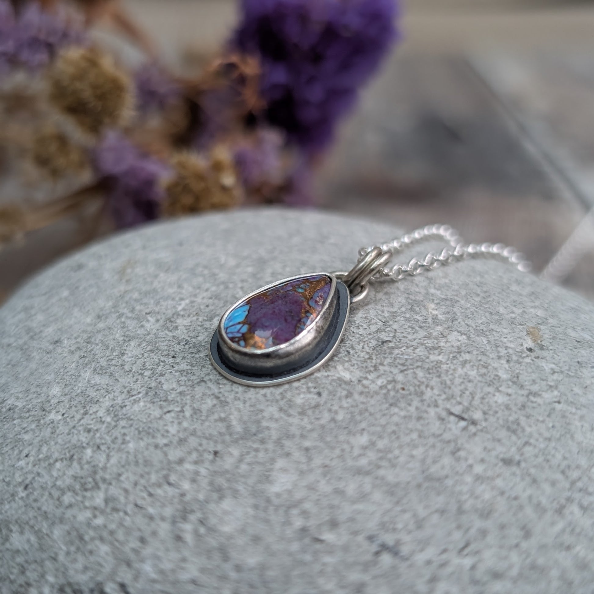 Close-up of the Mohave Turquoise necklace on a stone
Handmade recycled sterling silver necklace featuring a teardrop-shaped Mohave Turquoise stone with vibrant purple, blue, and copper tones. The oxidised silver setting contrasts with the bright silver chain, creating a unique, artisan jewellery piece.