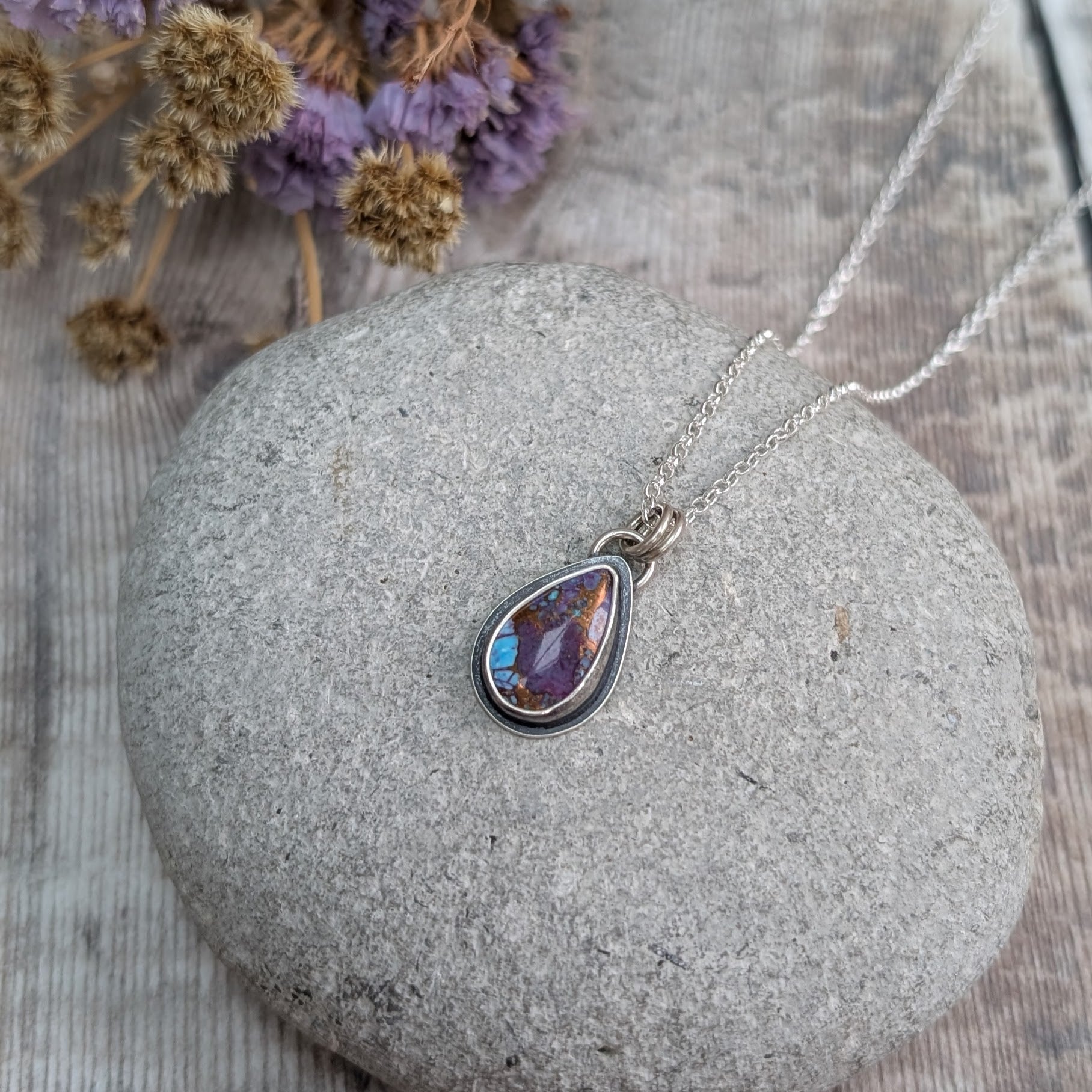 Close-up of the Mohave Turquoise necklace on a stone
Handmade recycled sterling silver necklace featuring a teardrop-shaped Mohave Turquoise stone with vibrant purple, blue, and copper tones. The oxidised silver setting contrasts with the bright silver chain, creating a unique, artisan jewellery piece.