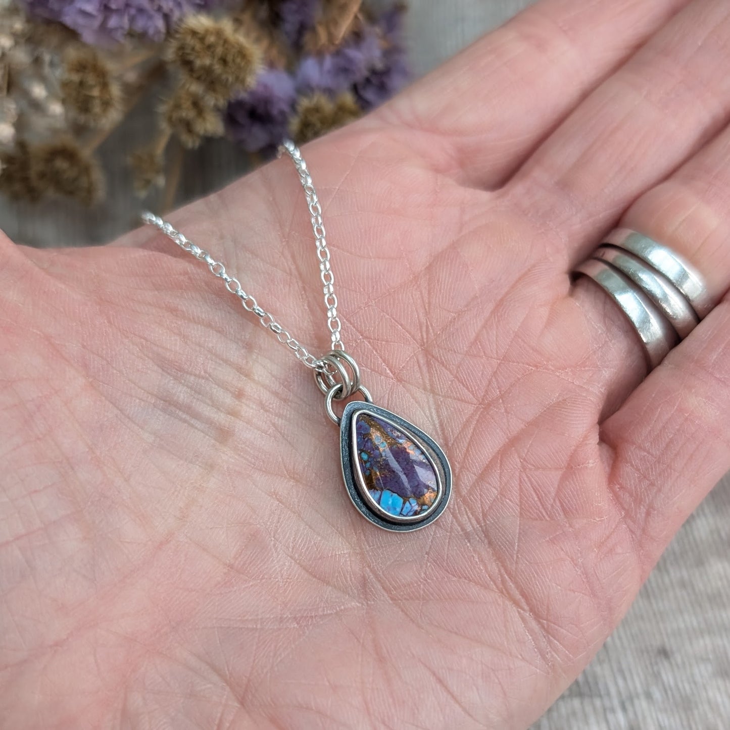 Mohave Turquoise necklace in hand for scale
Handmade Mohave Turquoise pendant necklace resting in a hand for scale. The teardrop gemstone, set in oxidised recycled sterling silver, showcases deep purple, blue, and copper hues. A delicate but striking artisan jewellery piece.