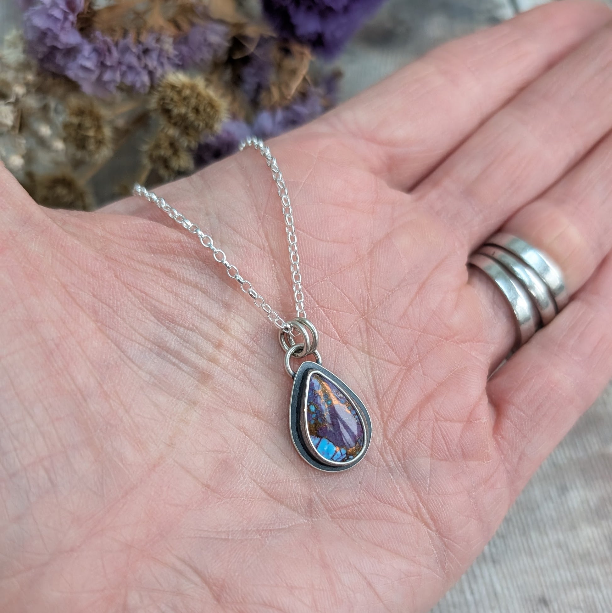 Mohave Turquoise necklace in hand for scale
Handmade Mohave Turquoise pendant necklace resting in a hand for scale. The teardrop gemstone, set in oxidised recycled sterling silver, showcases deep purple, blue, and copper hues. A delicate but striking artisan jewellery piece.