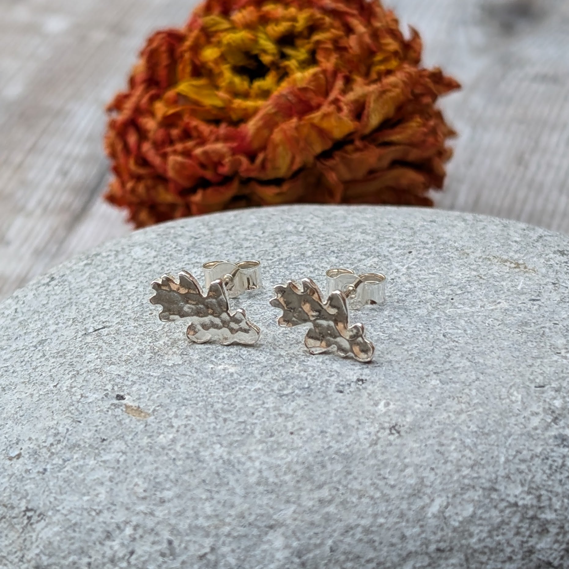 Close-up of a pair of sterling silver oak leaf stud earrings with a hammered finish, resting on a smooth grey stone with a blurred autumnal flower in the background.