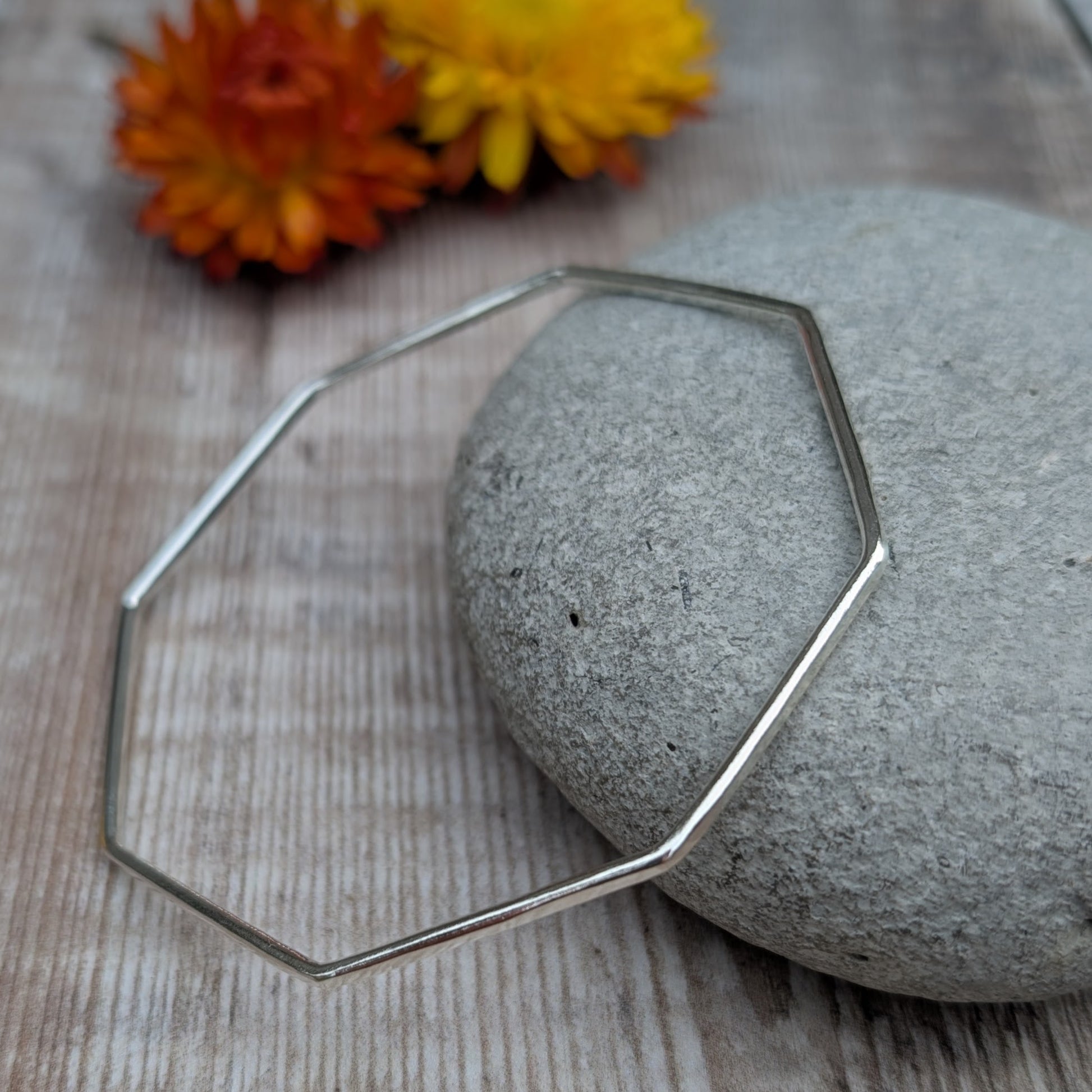 The geometric silver bangle displayed on a smooth stone with flowers in the background, emphasizing its minimalist and contemporary design