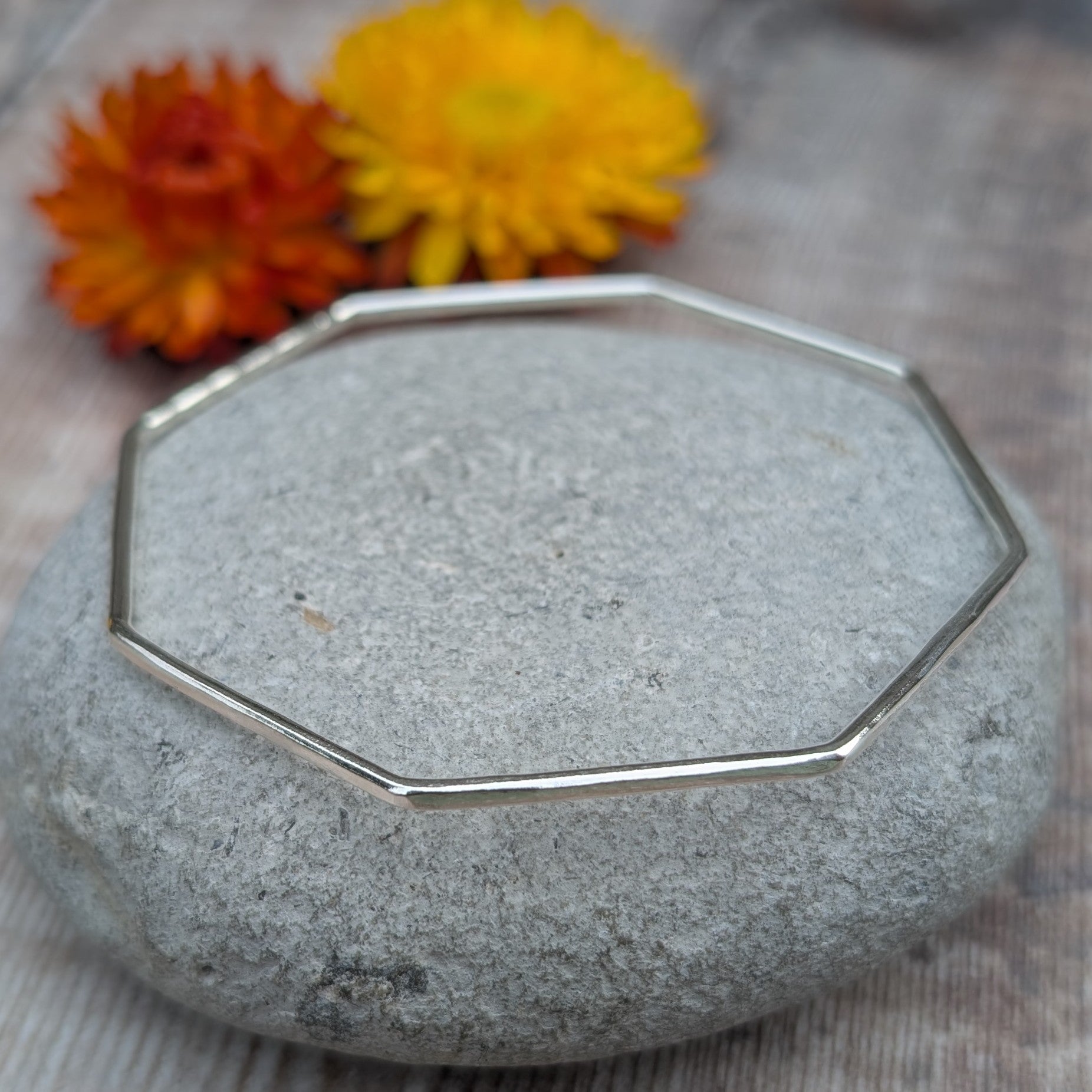The geometric silver bangle displayed on a smooth stone with flowers in the background, emphasizing its minimalist and contemporary design.