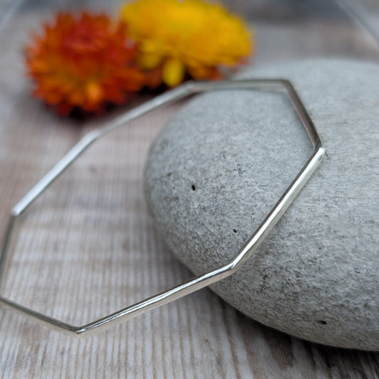 The geometric silver bangle displayed on a smooth stone with flowers in the background, emphasizing its minimalist and contemporary design.