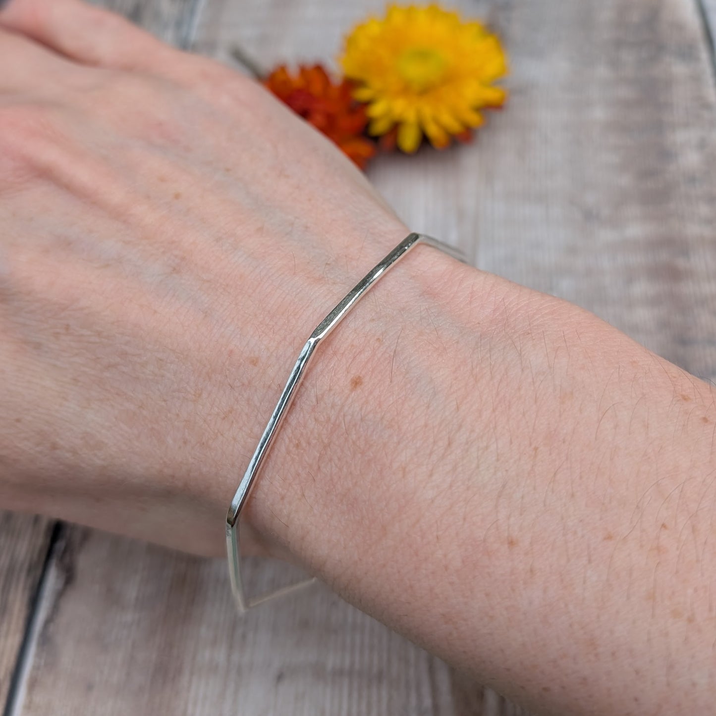 A recycled sterling silver octagon bangle worn on a wrist, showing its unique geometric shape against a rustic wooden background with yellow and orange flowers.