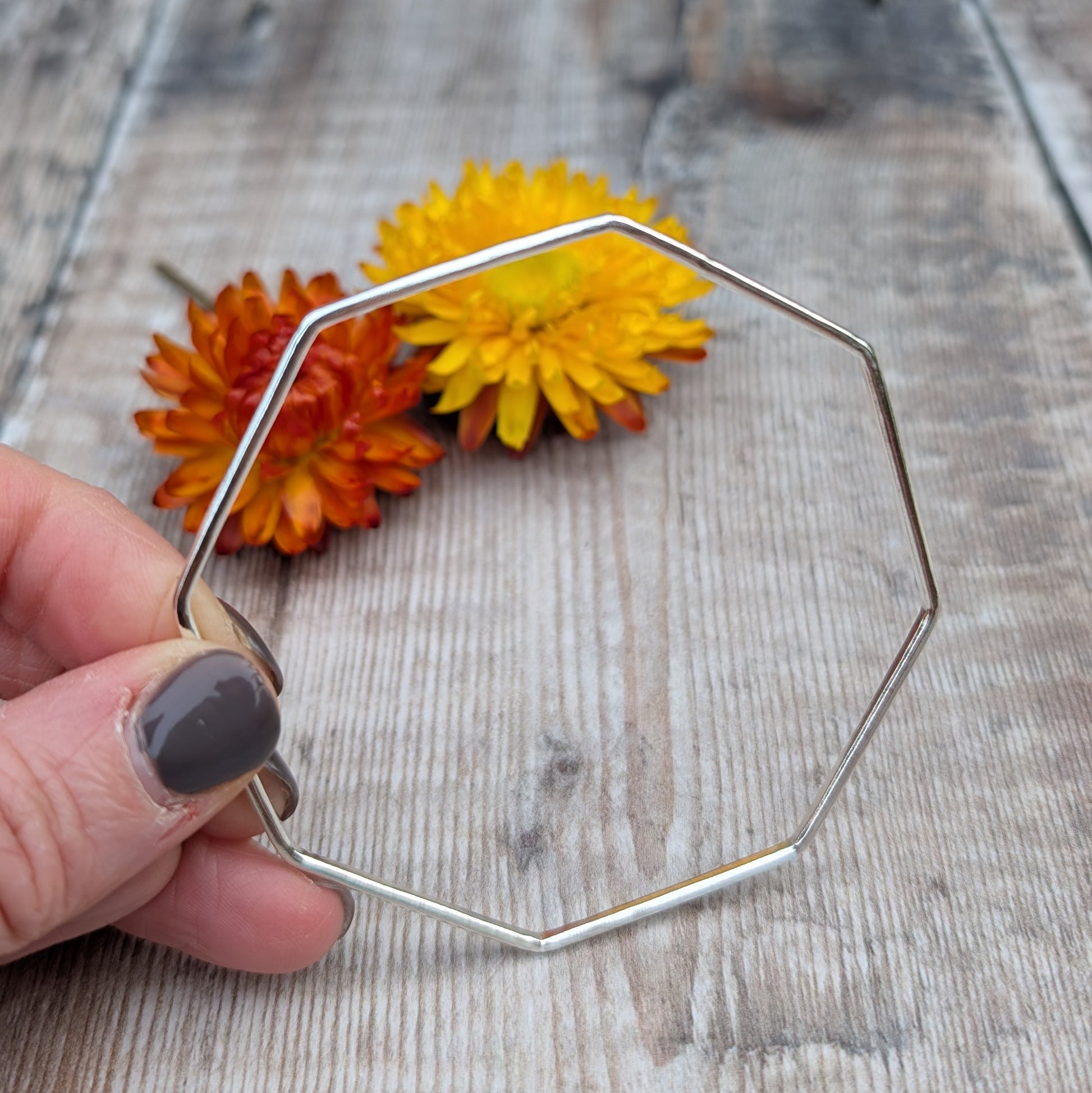 A close-up of the silver octagon bangle held between fingers, highlighting the smooth, angular design and polished finish.