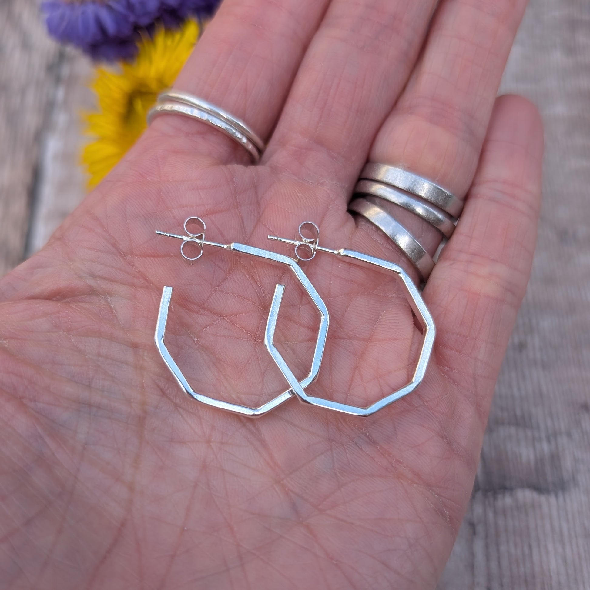 Close-up of handmade octagon hoop earrings in recycled sterling silver, displayed on an open hand with silver rings and a floral background.