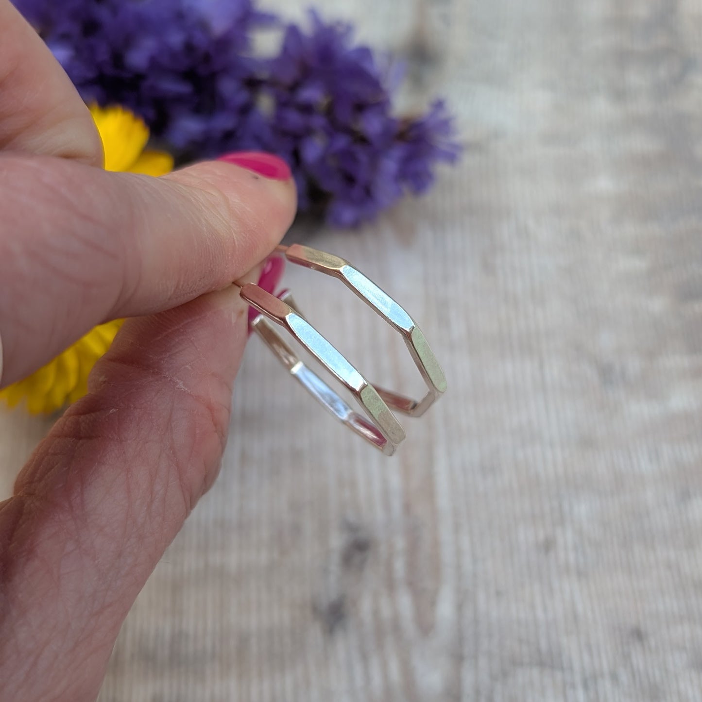 Side view of octagon hoop earrings, showcasing their faceted design, held between fingers against a blurred background of flowers.