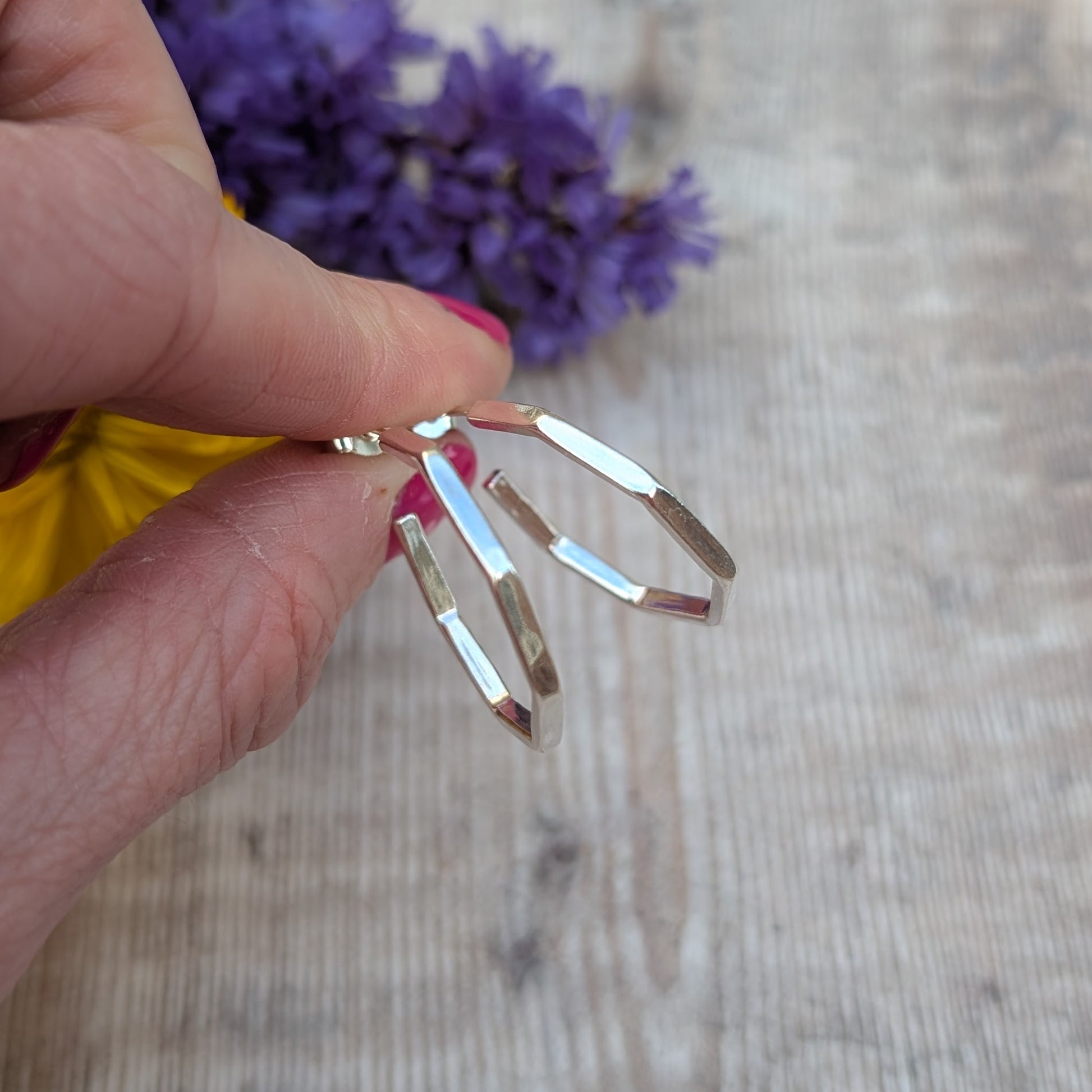 Side view of octagon hoop earrings, showcasing their faceted design, held between fingers against a blurred background of flowers.
