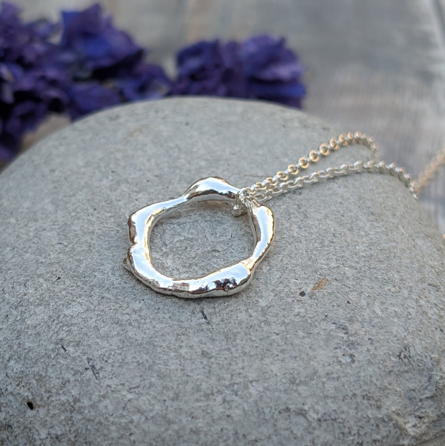A close-up of a handmade recycled sterling silver organic circle necklace resting on a smooth stone, with a blurred background of purple flowers.