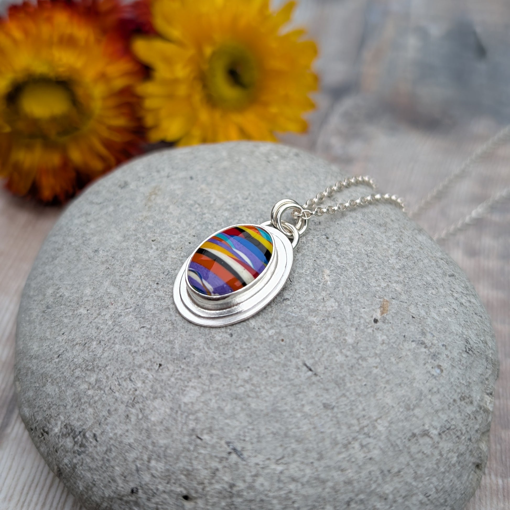 Handmade Surfite necklace displayed on a smooth stone with dried flowers in the background, showing the vibrant colours and layered detail of the pendant.