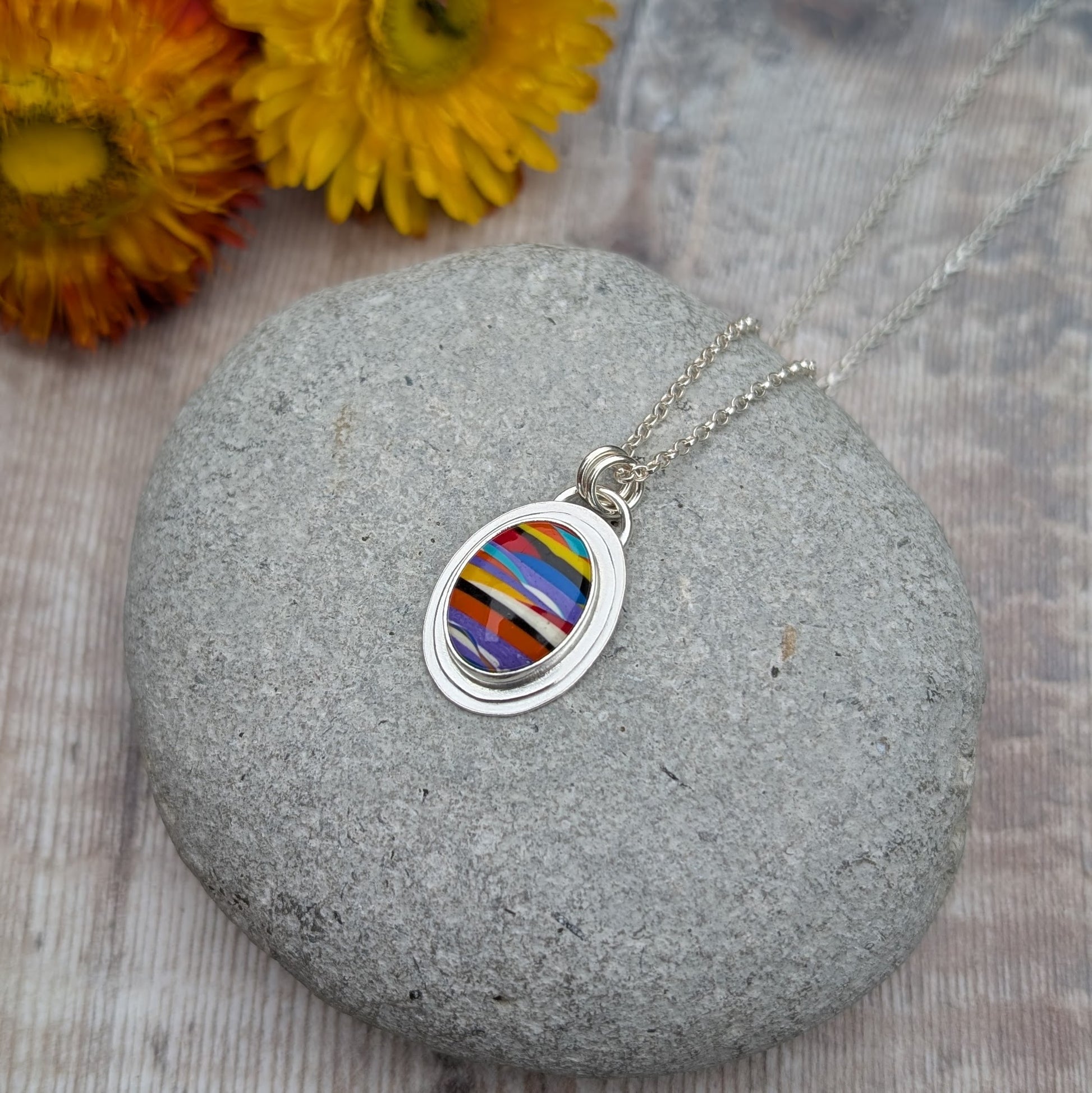 Handmade Surfite necklace displayed on a smooth stone with dried flowers in the background, showing the vibrant colours and layered detail of the pendant.