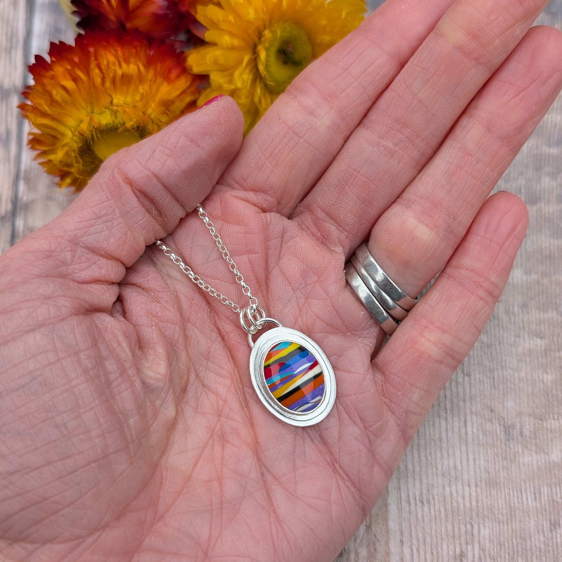 Close-up of a handmade oval Surfite necklace held in a hand, showing the colourful layered Surfite stone set in a sterling silver pendant.