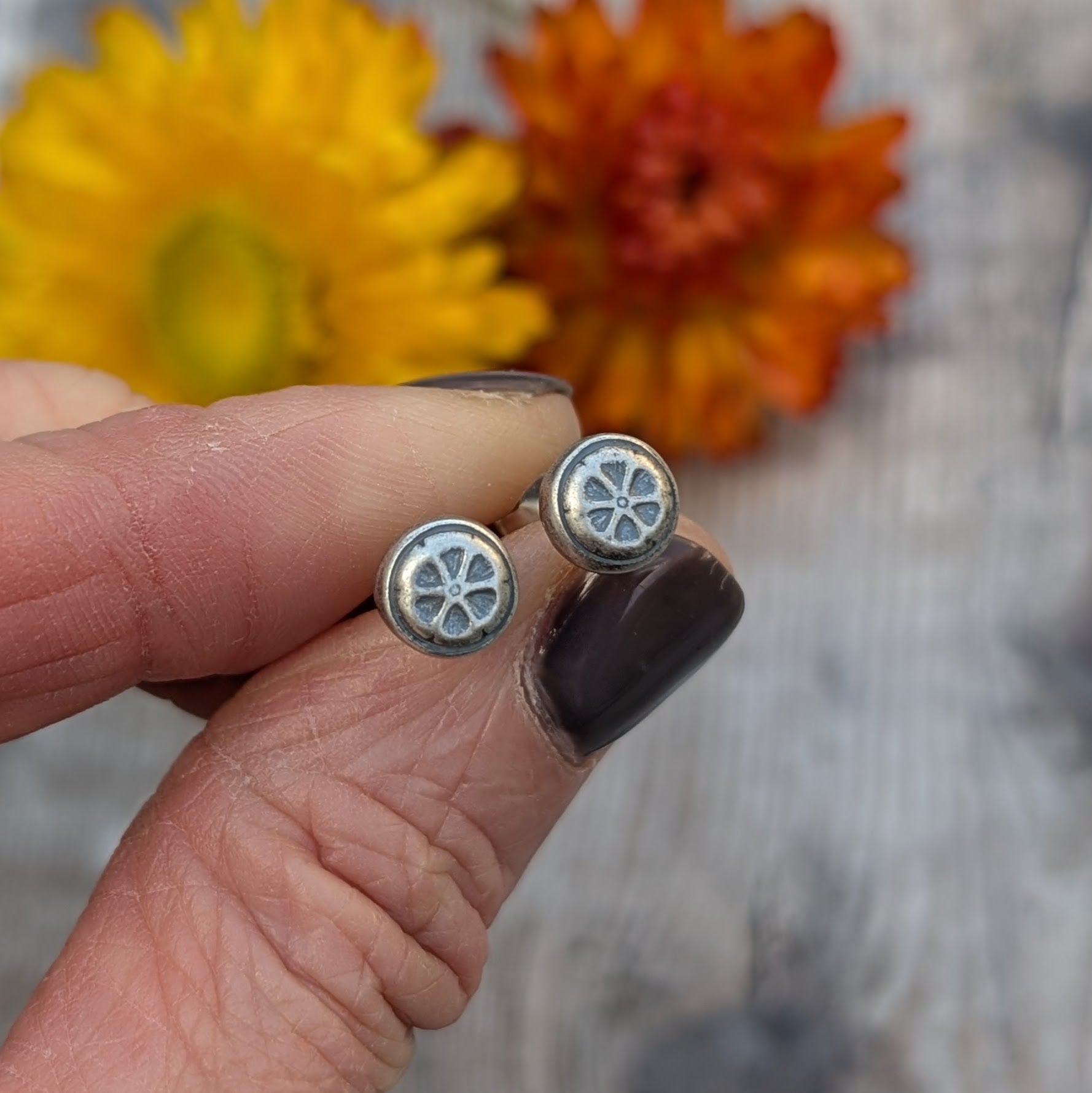 Hand holding the silver flower stud earrings, showing the oxidised details against a background of dried yellow and orange flowers.