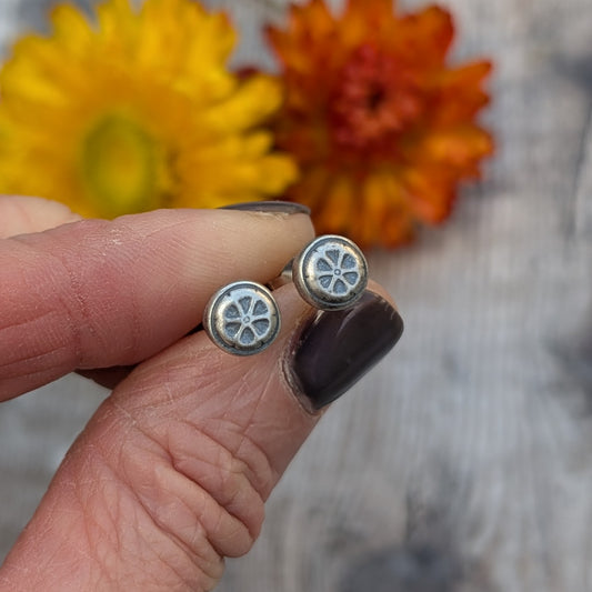 Hand holding the silver flower stud earrings, showing the oxidised details against a background of dried yellow and orange flowers.