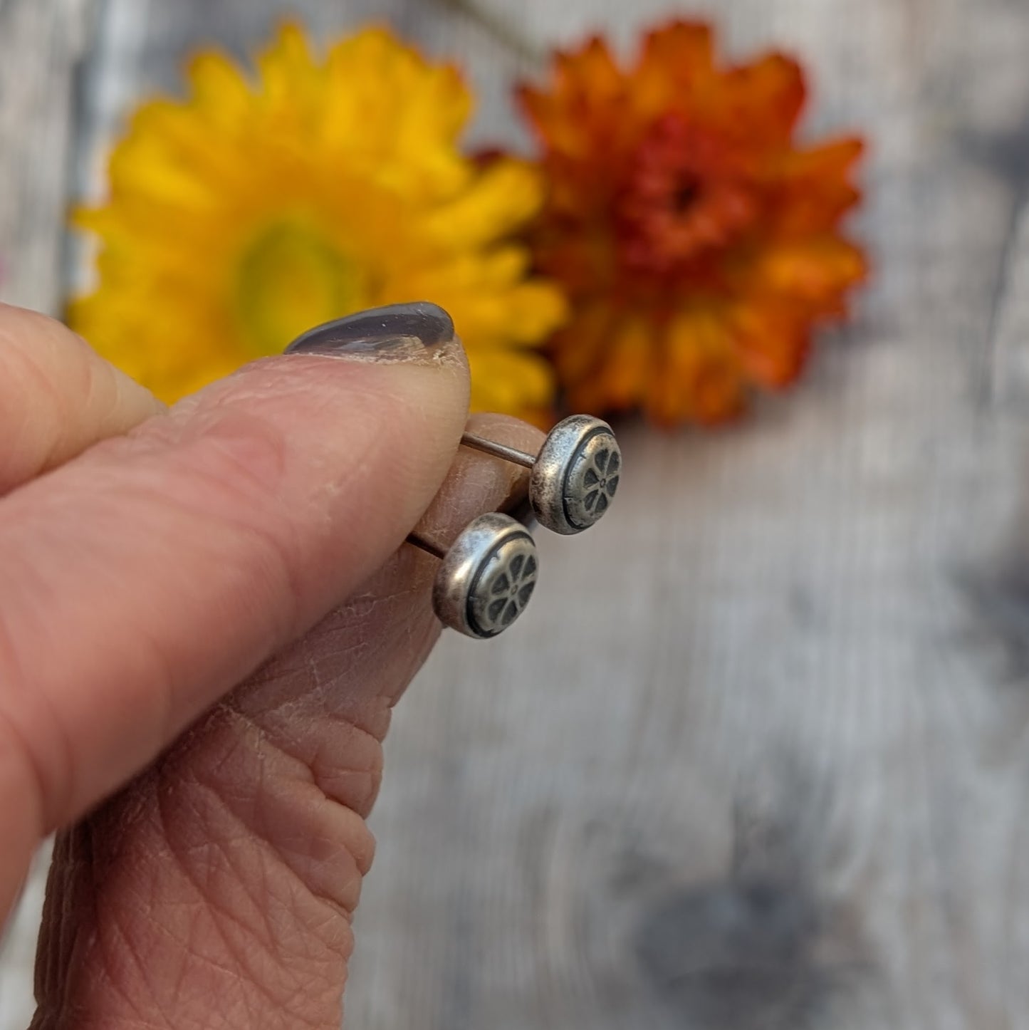 Hand holding the silver flower stud earrings, showing the oxidised details against a background of dried yellow and orange flowers.