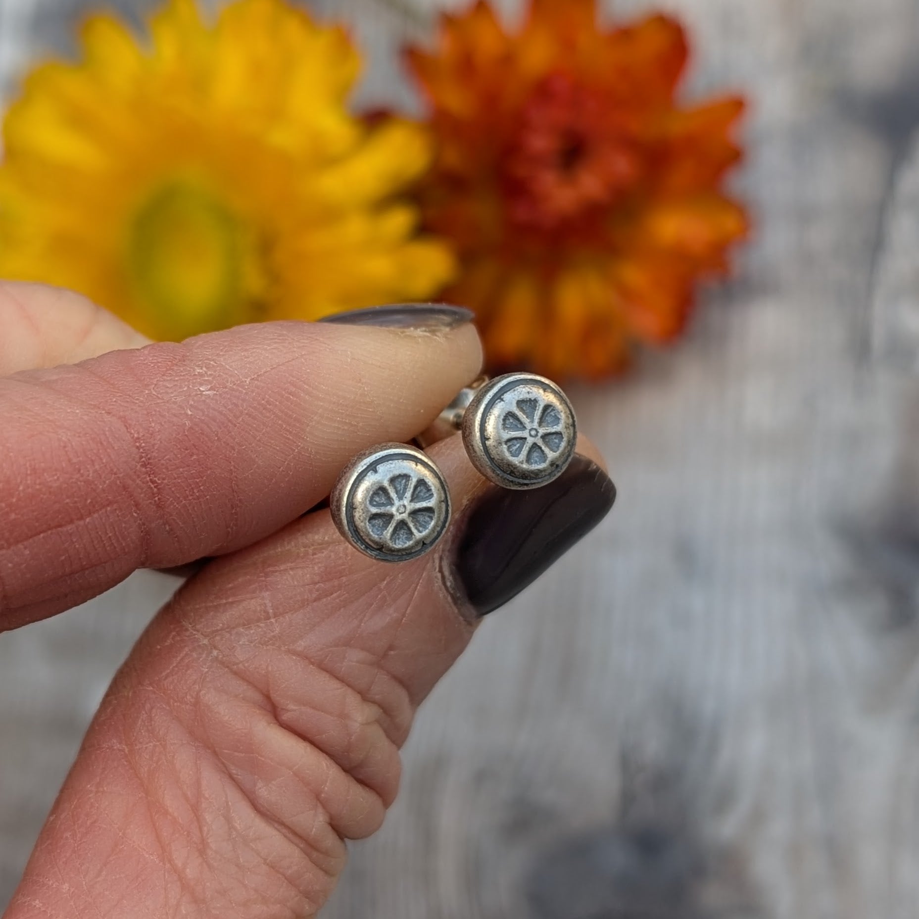 Hand holding the silver flower stud earrings, showing the oxidised details against a background of dried yellow and orange flowers.