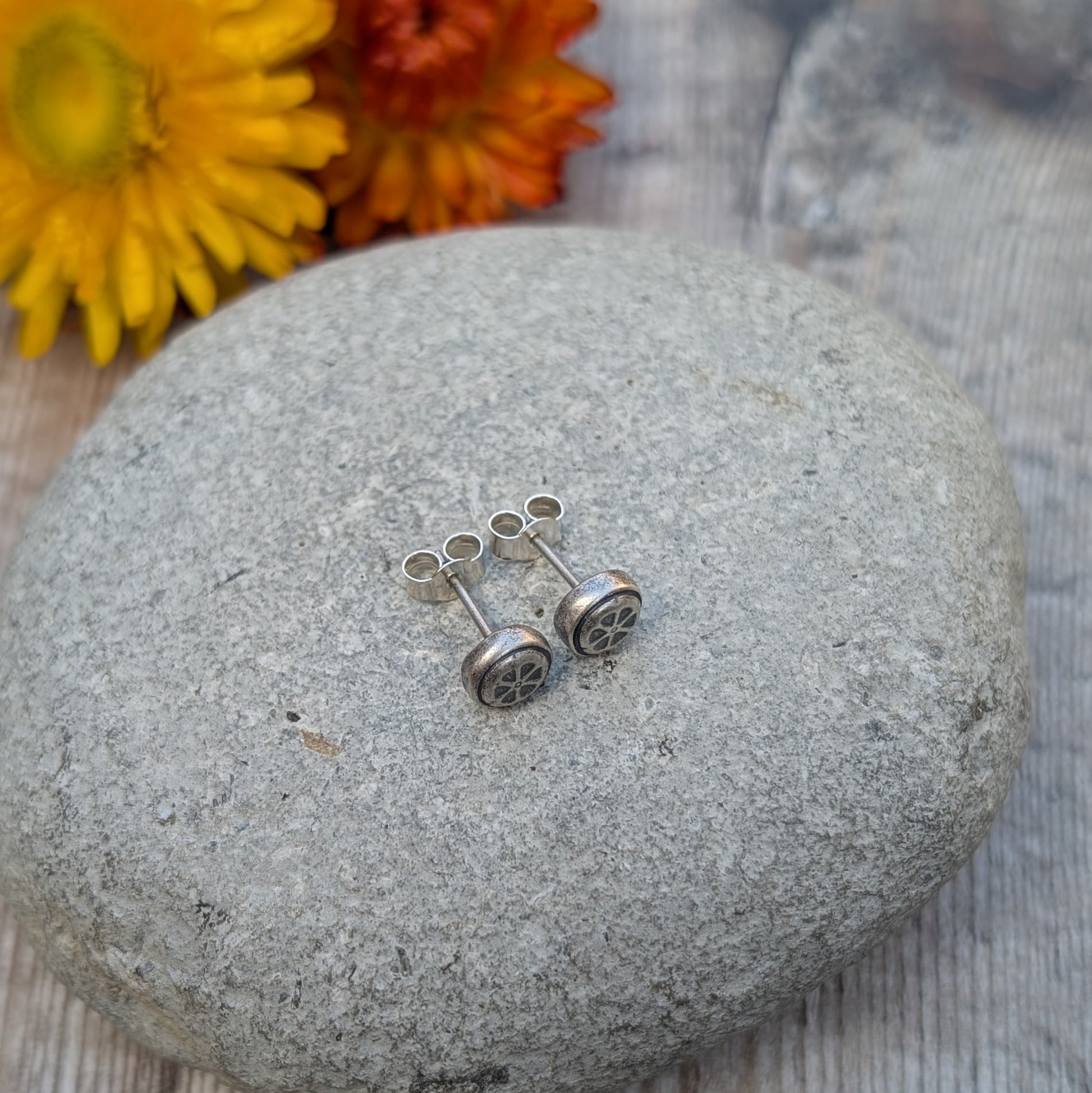 A pair of recycled sterling silver stud earrings with a pressed flower design, placed on a smooth stone with yellow and orange flowers in the background.
