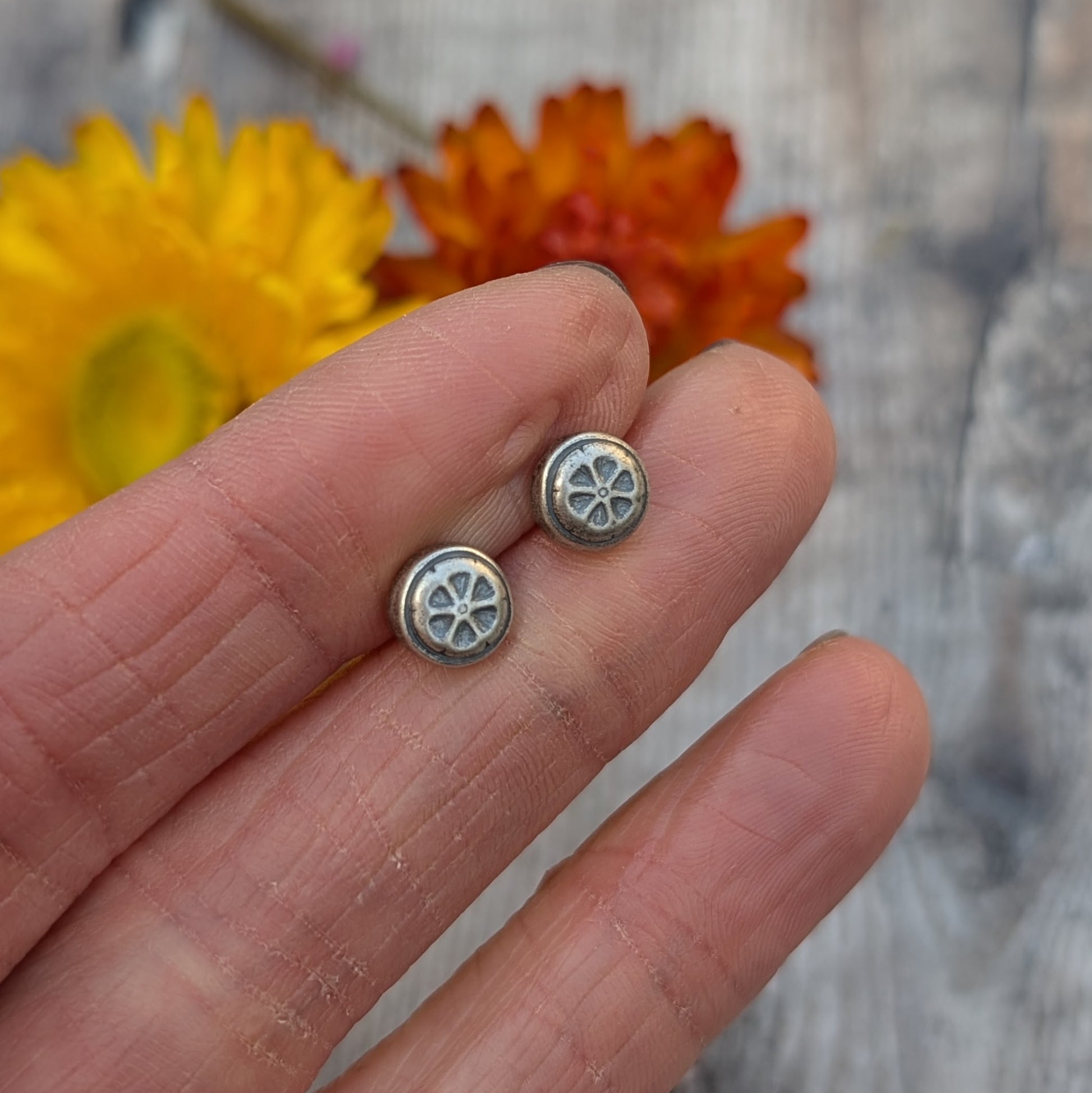 Hand holding the silver flower stud earrings, showing the oxidised details against a background of dried yellow and orange flowers.