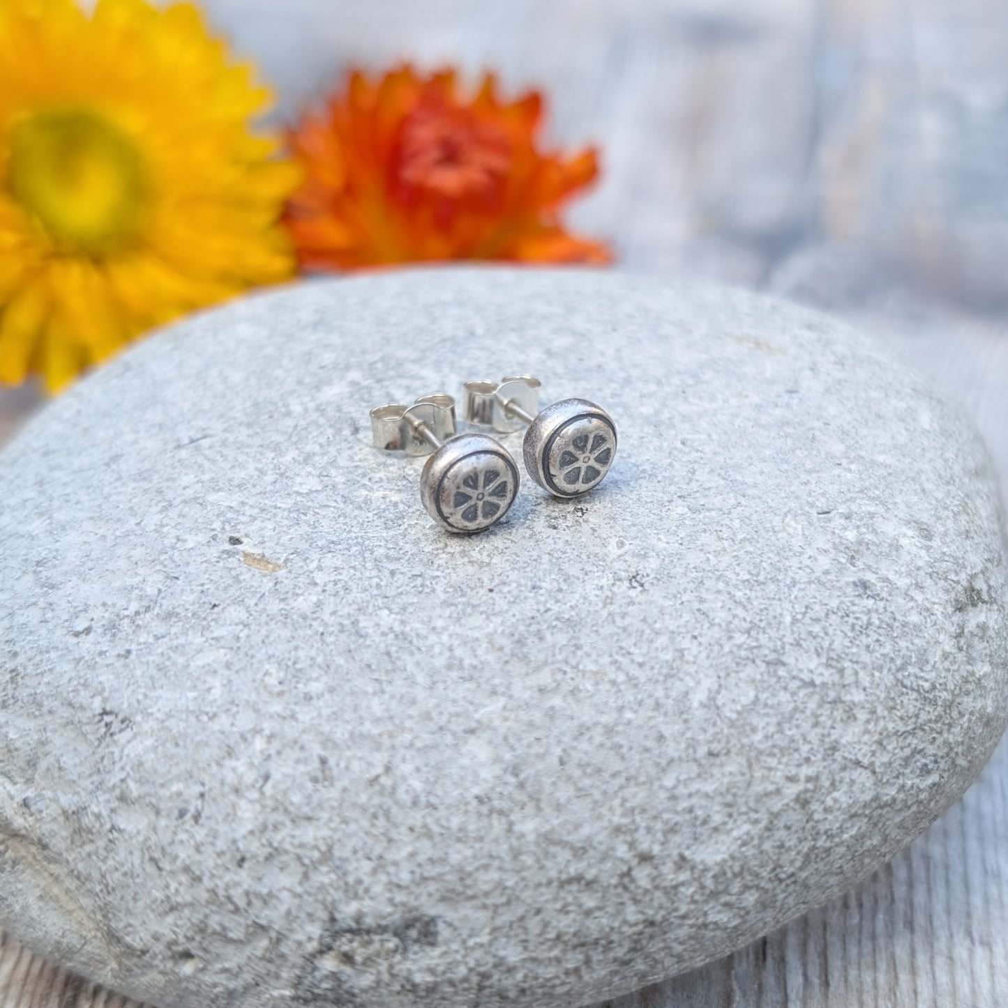 A pair of recycled sterling silver stud earrings with a pressed flower design, placed on a smooth stone with yellow and orange flowers in the background.
