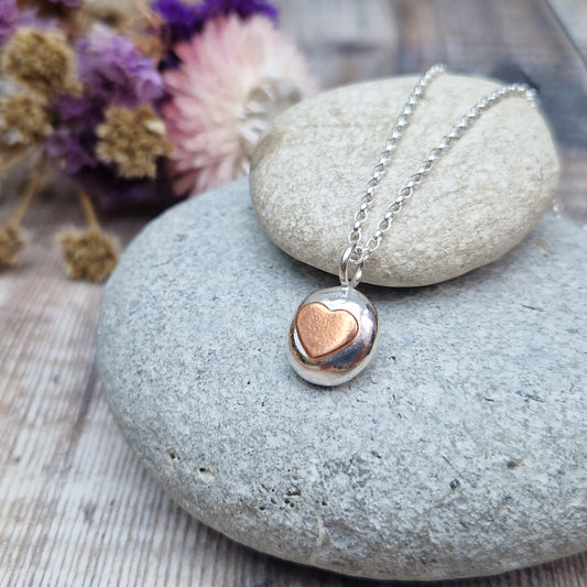 Sterling Silver round pebble shaped pendant with slightly raised silver heart shape at its centre. Suspended from silver chain via small circle link.