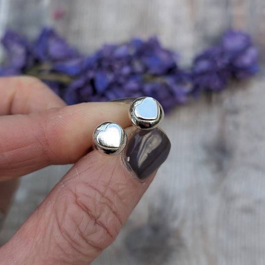 A hand holding the silver heart stud earrings, highlighting their organic shape and reflective surface against a blurred floral background.