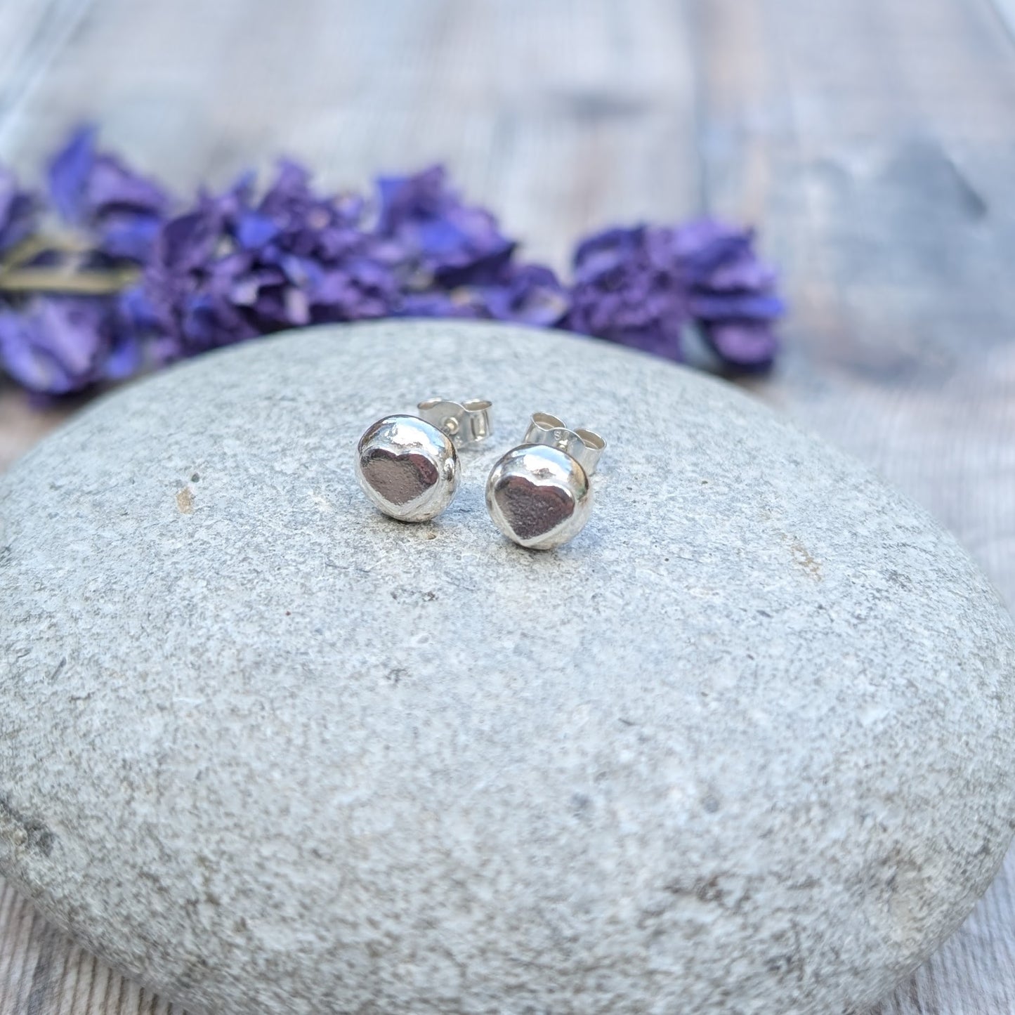 A pair of handmade sterling silver heart stud earrings resting on a smooth stone, with purple flowers in the background.