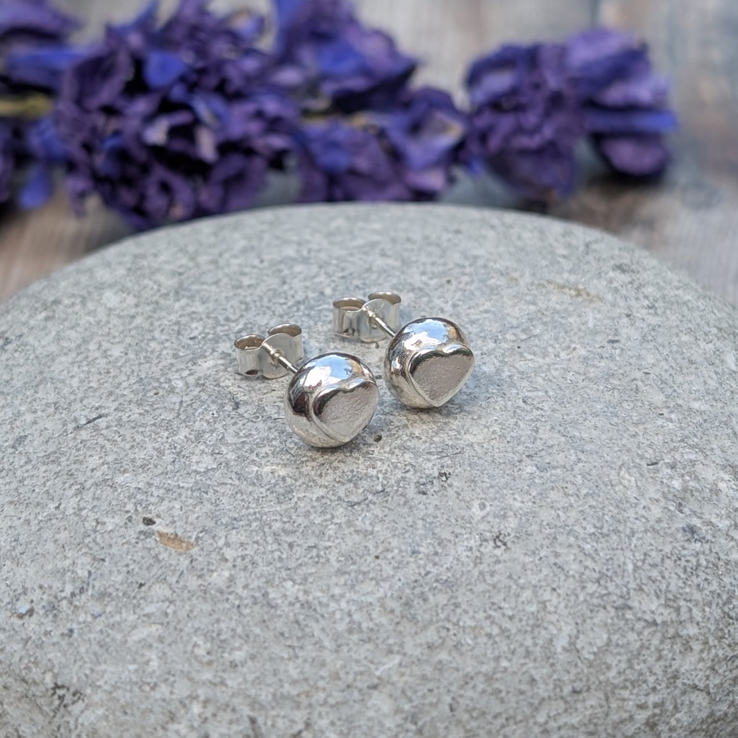 A pair of handmade sterling silver heart stud earrings resting on a smooth stone, with purple flowers in the background.