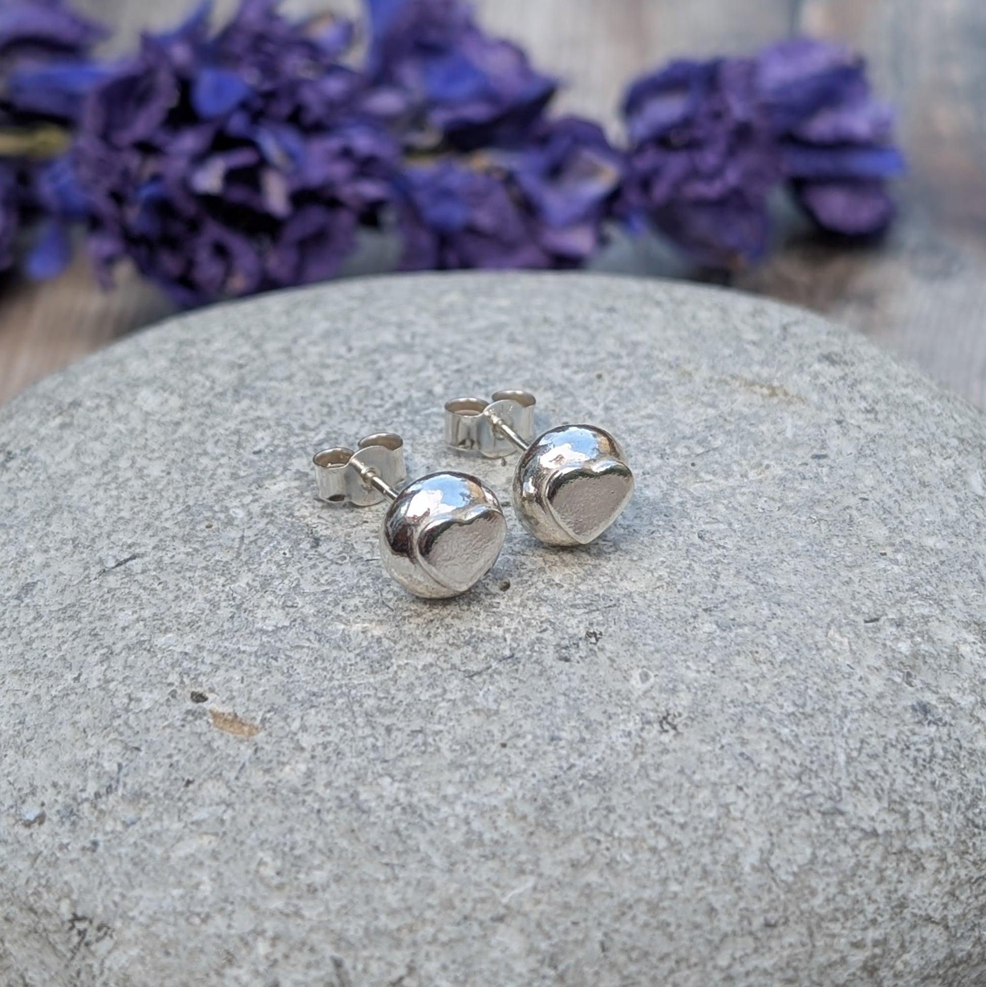 A pair of handmade sterling silver heart stud earrings resting on a smooth stone, with purple flowers in the background.