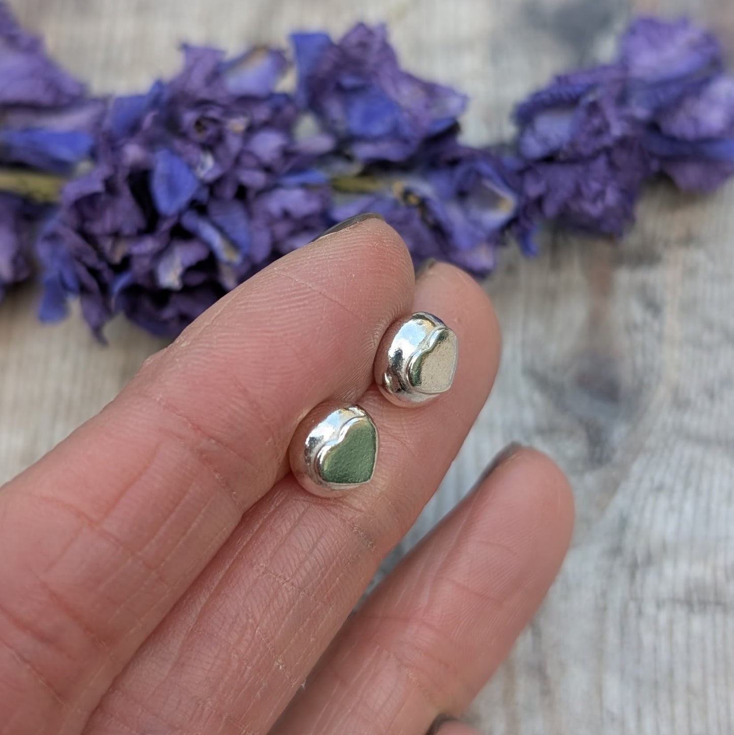 A hand holding the silver heart stud earrings, highlighting their organic shape and reflective surface against a blurred floral background.