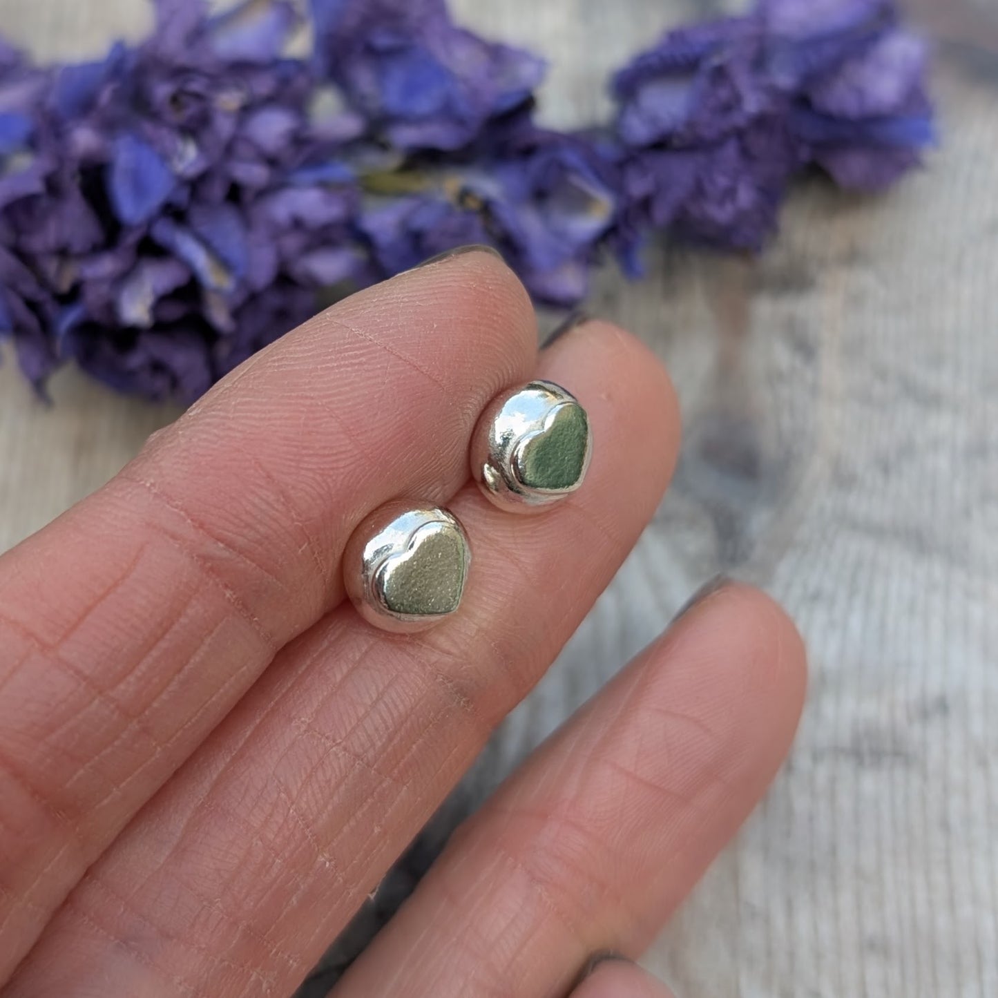 A hand holding the silver heart stud earrings, highlighting their organic shape and reflective surface against a blurred floral background.