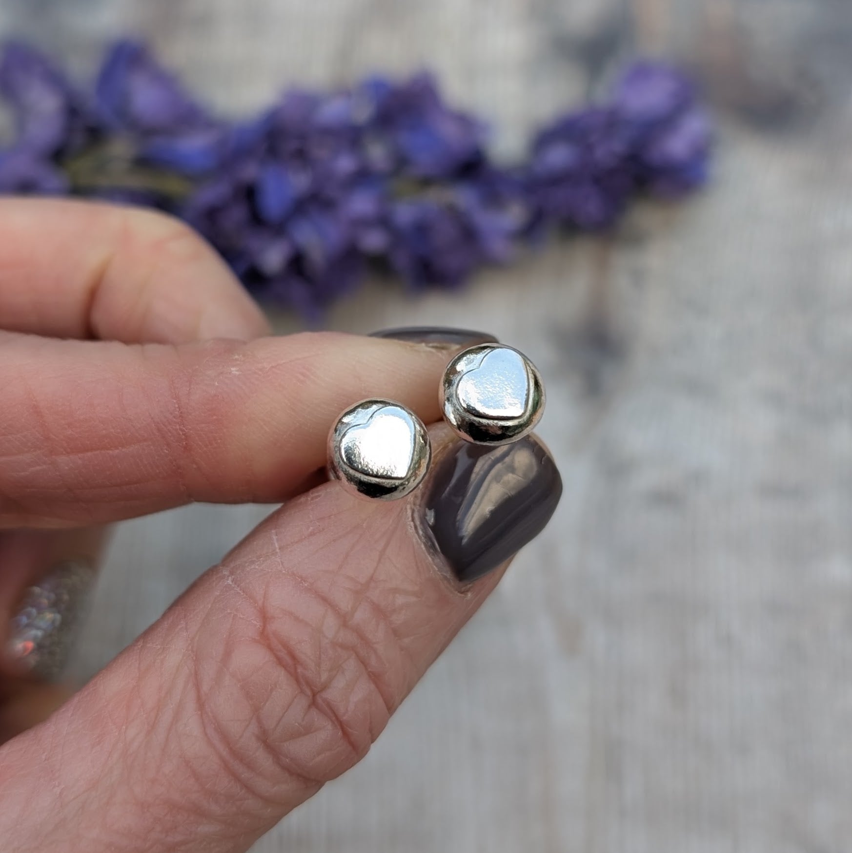 A hand holding the silver heart stud earrings, highlighting their organic shape and reflective surface against a blurred floral background.