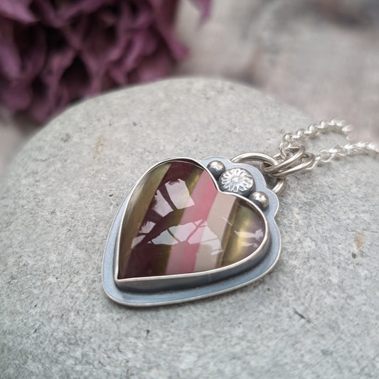 Pendant is a heart shape surfite stone with smooth gloss finish. Resin stripes run diagonally light pink, pink, gold, brown and light brown. The oxidised silver bezel surround has a raised floral detail above the mounted stone and a small, raised sphere on each side. The pendant is attached to a silver chain via a ‘D’ link and two small open circle links.