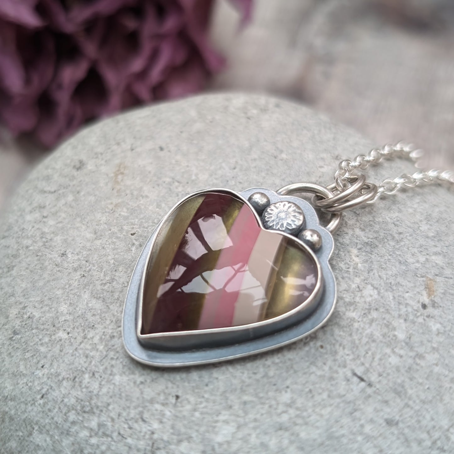 Pendant is a heart shape surfite stone with smooth gloss finish. Resin stripes run diagonally light pink, pink, gold, brown and light brown. The oxidised silver bezel surround has a raised floral detail above the mounted stone and a small, raised sphere on each side. The pendant is attached to a silver chain via a ‘D’ link and two small open circle links.