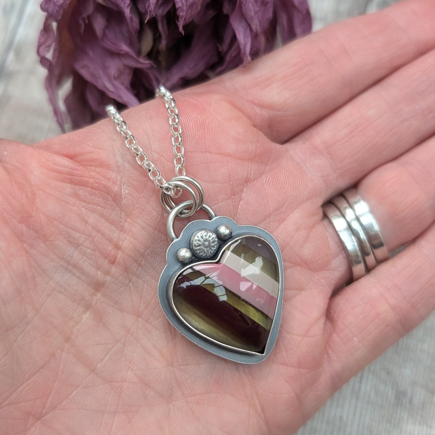 Pendant is a heart shape surfite stone with smooth gloss finish. Resin stripes run diagonally light pink, pink, gold, brown and light brown. The oxidised silver bezel surround has a raised floral detail above the mounted stone and a small, raised sphere on each side. The pendant is attached to a silver chain via a ‘D’ link and two small open circle links.
