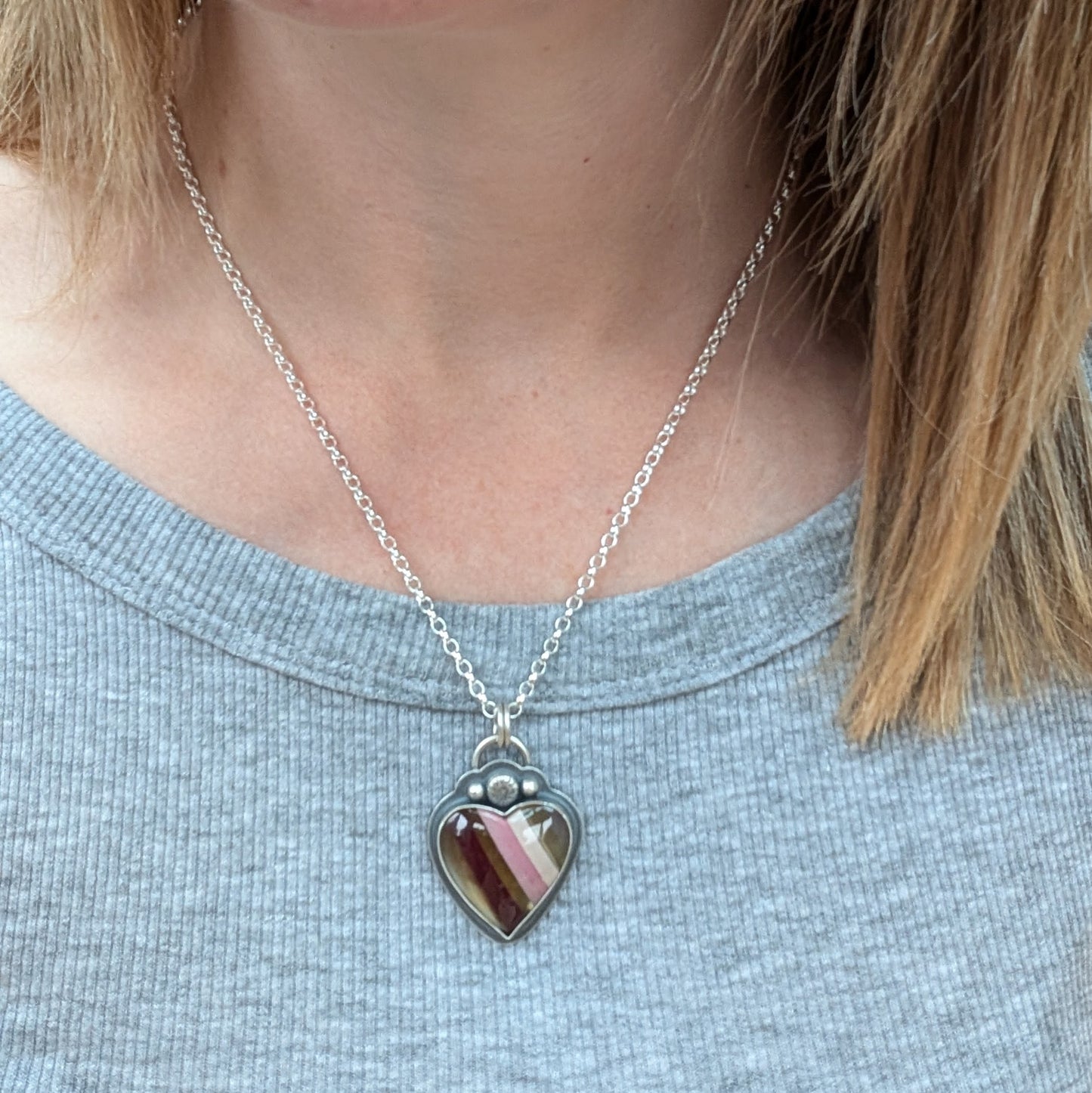 Pendant is a heart shape surfite stone with smooth gloss finish. Resin stripes run diagonally light pink, pink, gold, brown and light brown. The oxidised silver bezel surround has a raised floral detail above the mounted stone and a small, raised sphere on each side. The pendant is attached to a silver chain via a ‘D’ link and two small open circle links.