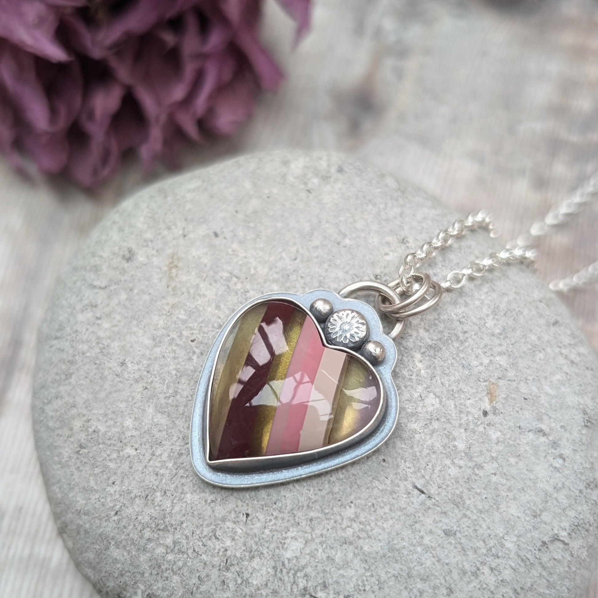 Pendant is a heart shape surfite stone with smooth gloss finish. Resin stripes run diagonally light pink, pink, gold, brown and light brown. The oxidised silver bezel surround has a raised floral detail above the mounted stone and a small, raised sphere on each side. The pendant is attached to a silver chain via a ‘D’ link and two small open circle links.