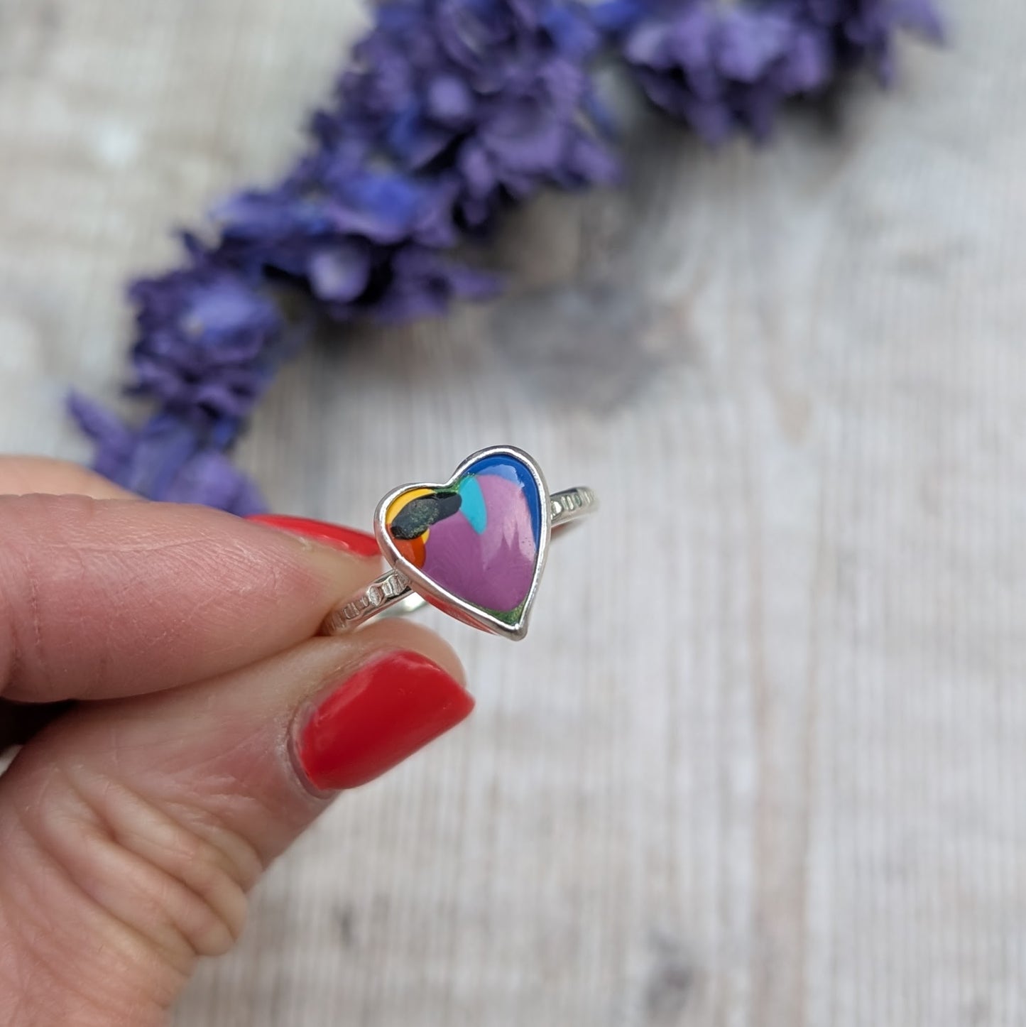 Close-up of the heart-shaped Surfite ring being held in fingers, with a background of dried purple flowers.