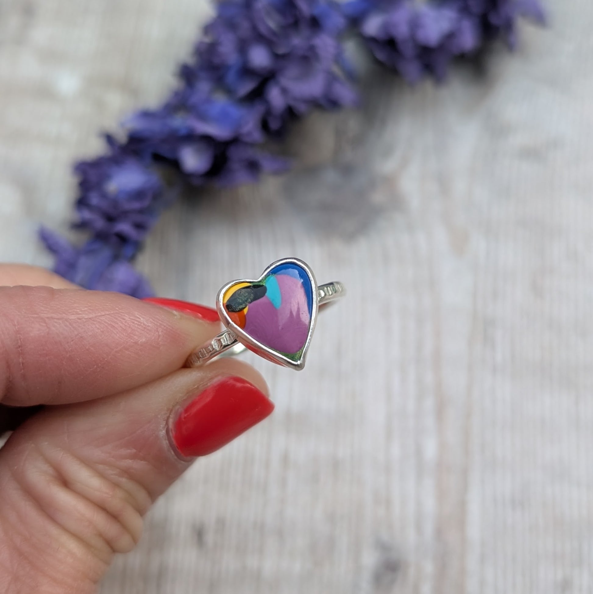 Close-up of the heart-shaped Surfite ring being held in fingers, with a background of dried purple flowers.