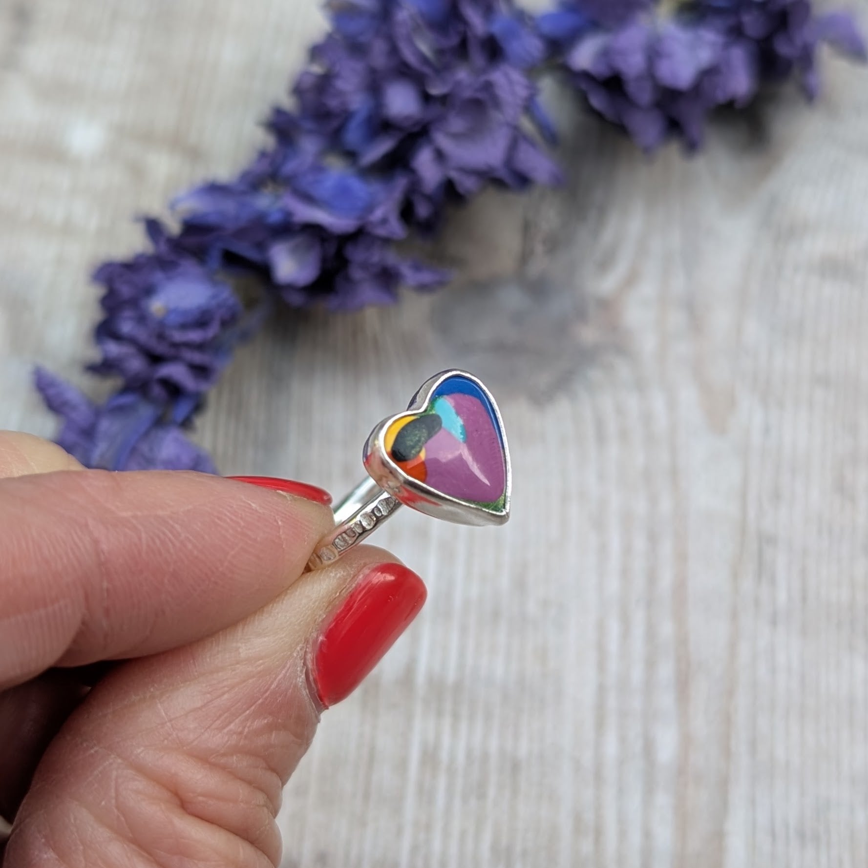 Close-up of the heart-shaped Surfite ring being held in fingers, with a background of dried purple flowers.