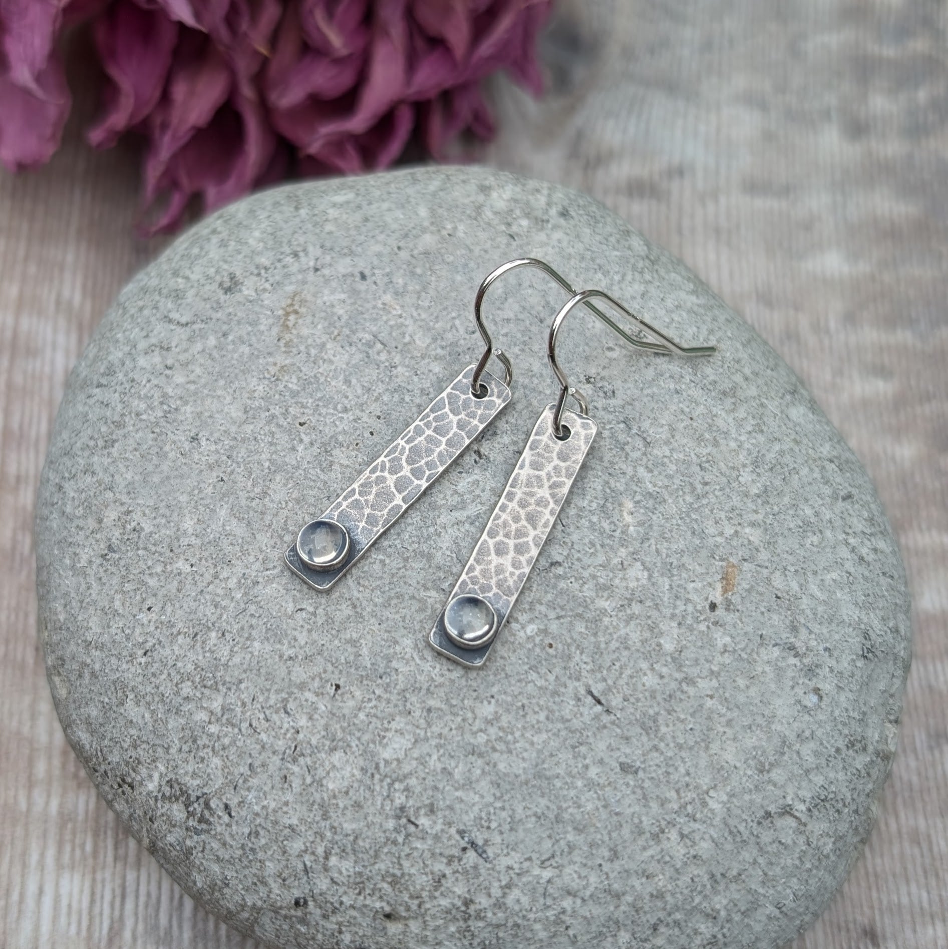 Pair of oxidised sterling silver rectangle earrings with a hammered texture, placed on a smooth stone, each earring featuring a moonstone at the base.