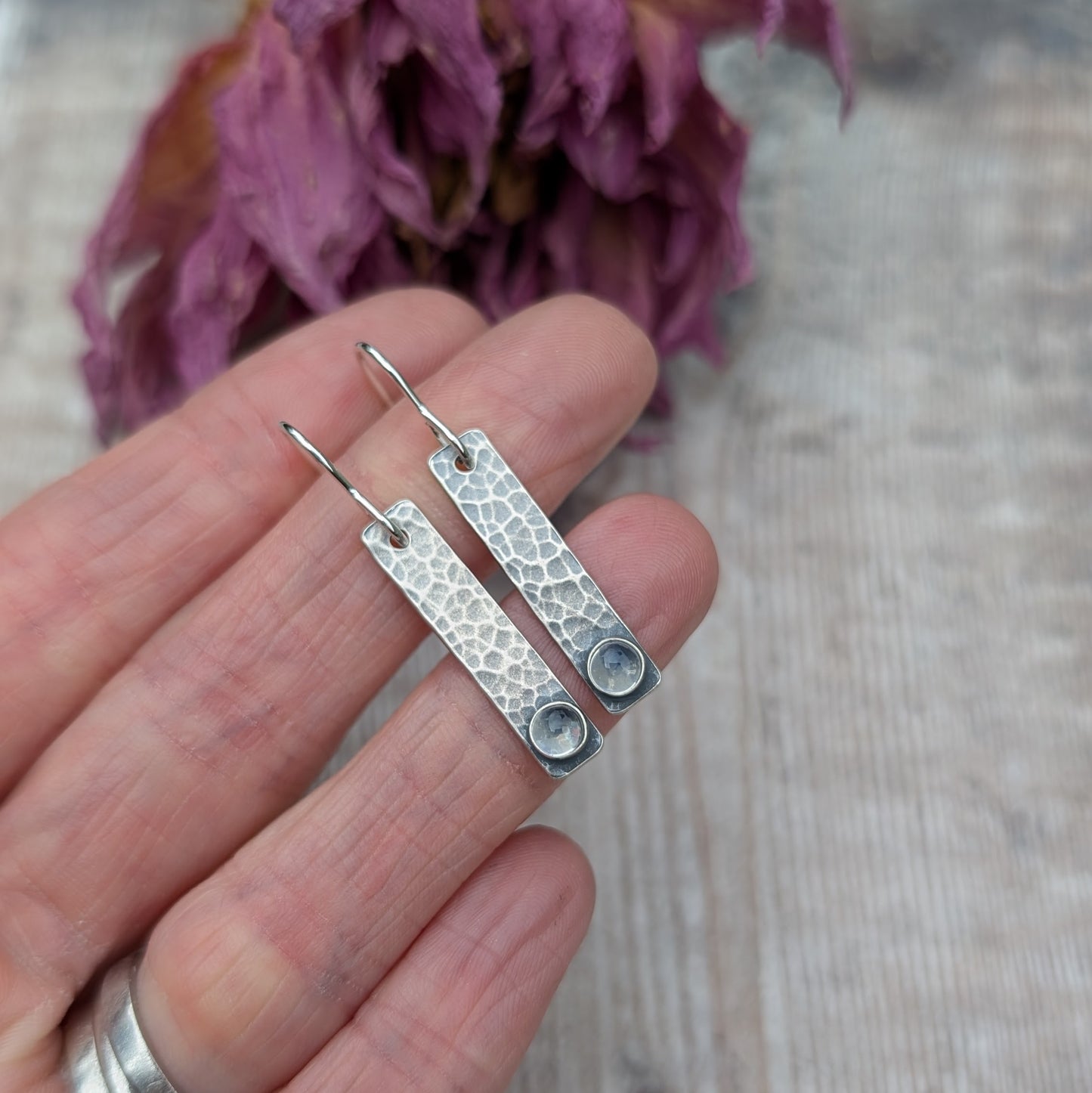 Close-up of a hand holding a pair of oxidised sterling silver rectangle earrings with a hammered texture, featuring a moonstone at the base of each earring.