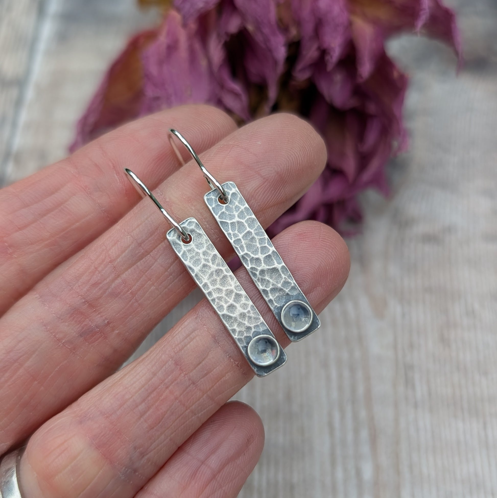 Close-up of a hand holding a pair of oxidised sterling silver rectangle earrings with a hammered texture, featuring a moonstone at the base of each earring.