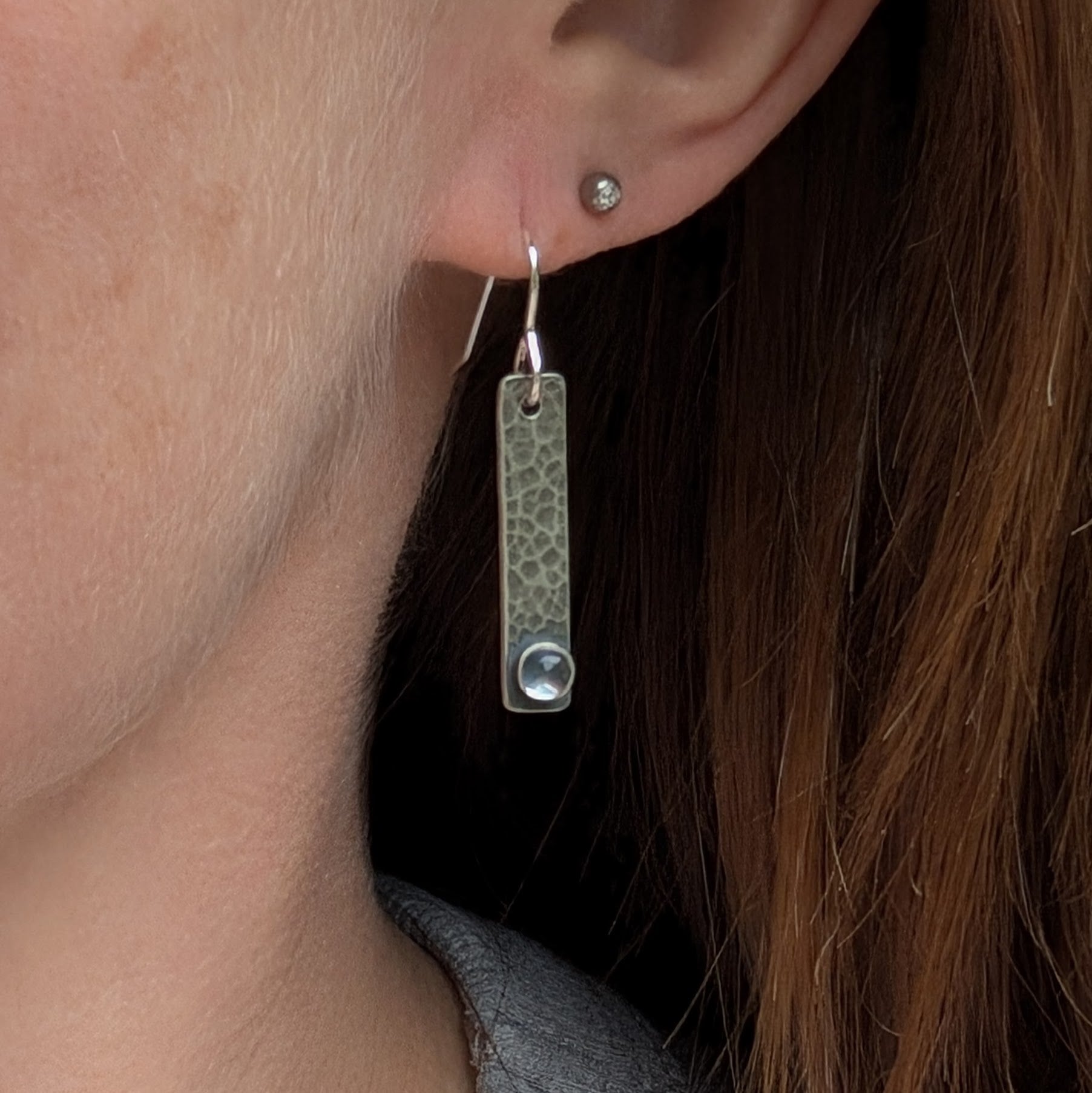 Side view of a person wearing a single oxidised sterling silver rectangle earring with a hammered texture and a moonstone accent at the base.