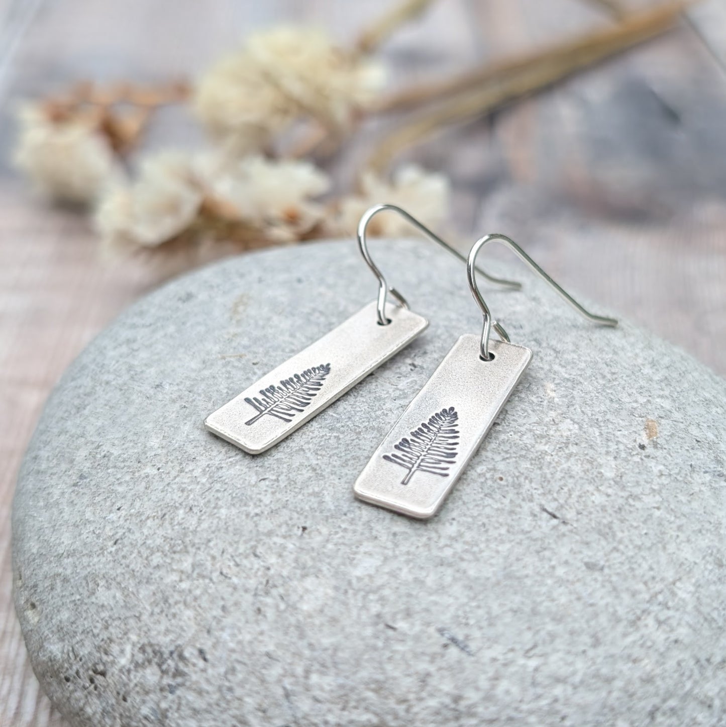 A pair of oxidised silver rectangle earrings featuring an etched evergreen tree design, displayed on a smooth grey stone. Softly blurred dried flowers in the background enhance the natural theme.