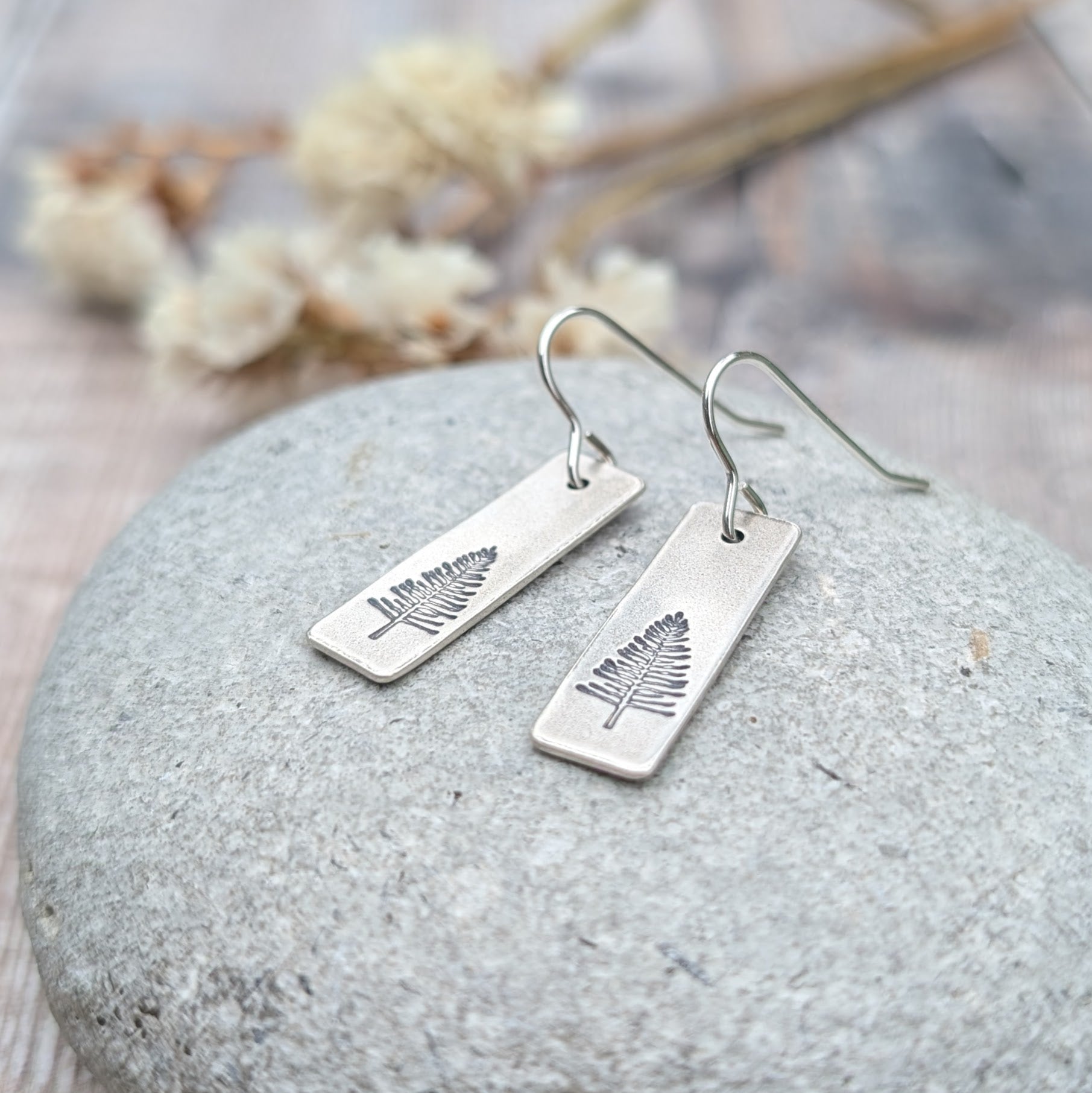A pair of oxidised silver rectangle earrings featuring an etched evergreen tree design, displayed on a smooth grey stone. Softly blurred dried flowers in the background enhance the natural theme.